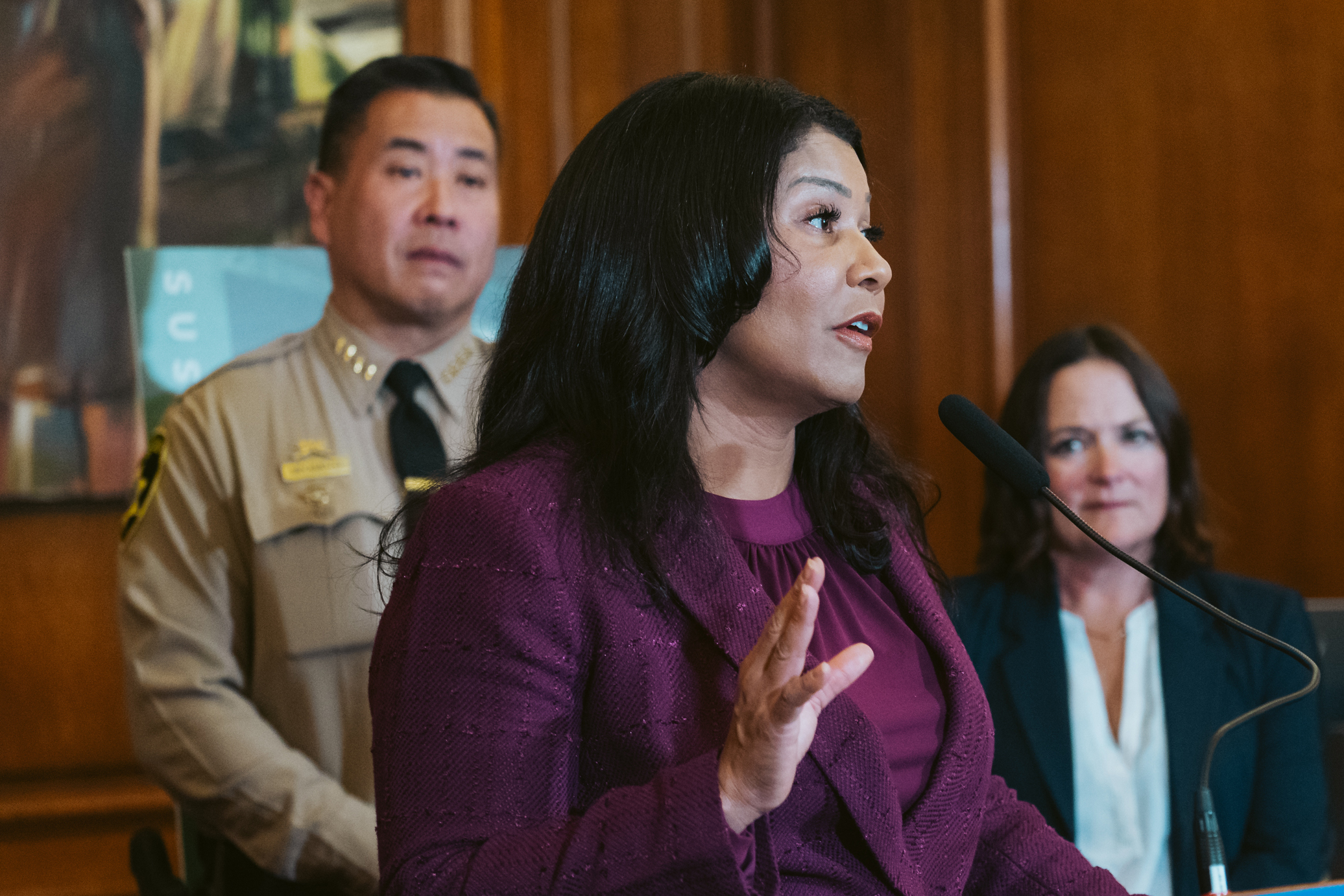 San Francisco Mayor London Breed, dressed in a purple speaks to the media during a press conference in City Hall. She sands behind a podium that reads &quot;APEC 2023 UNITED STATES&quot; in a wood paneled room.