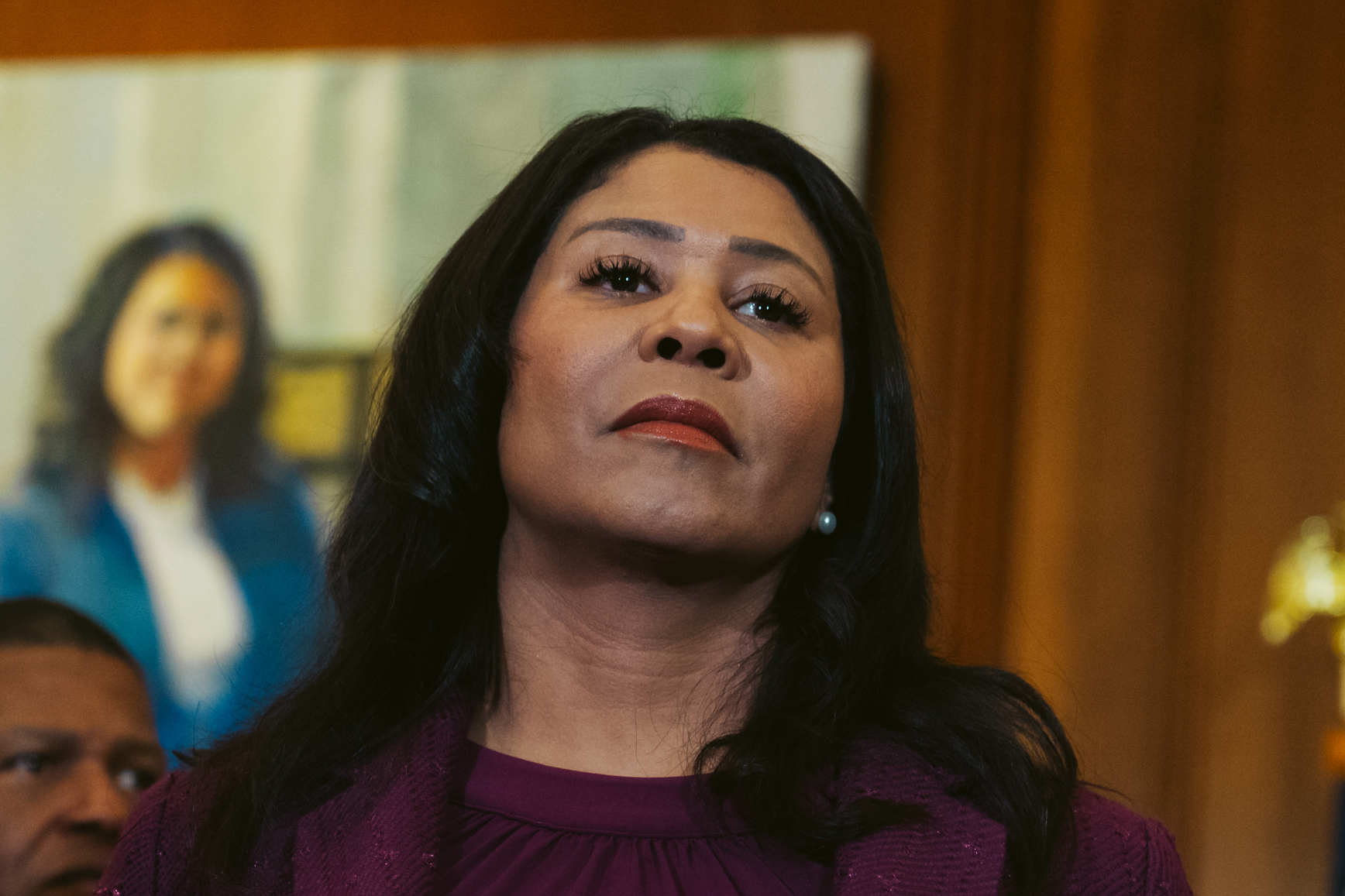 San Francisco Mayor London Breed, dressed in a purple speaks to the media during a press conference in City Hall. She stands in a wood paneled room with the American flag in the background.
