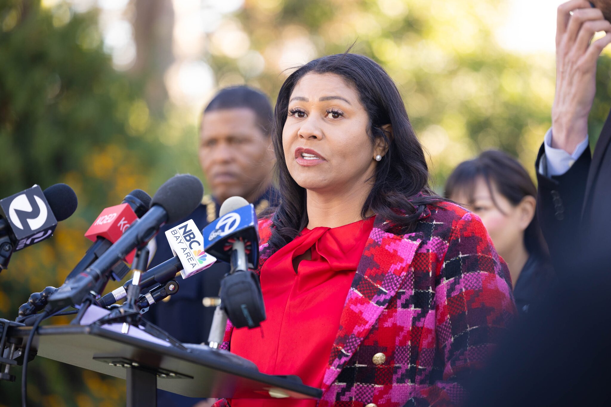 A woman in a red shirt and plaid blazer speaks into a microphone.
