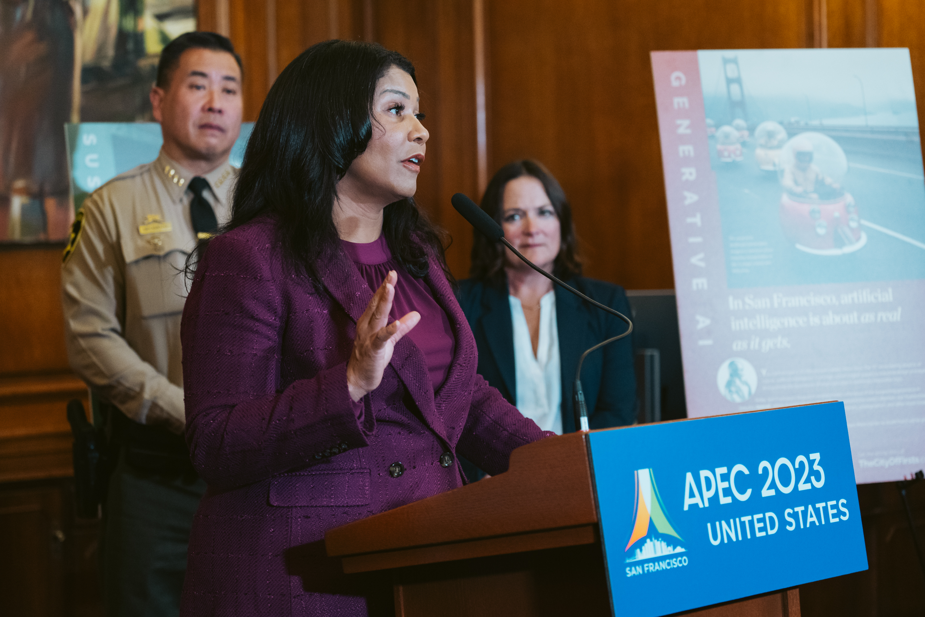 San Francisco Mayor London Breed, dressed in a purple suit raises her right hand while gesticulating as she speaks to the media during a press conference in City Hall. She sands behind a podium that reads &quot;APEC 2023 UNITED STATES&quot; in a wood paneled room with two other people standing behind her listening attentively.