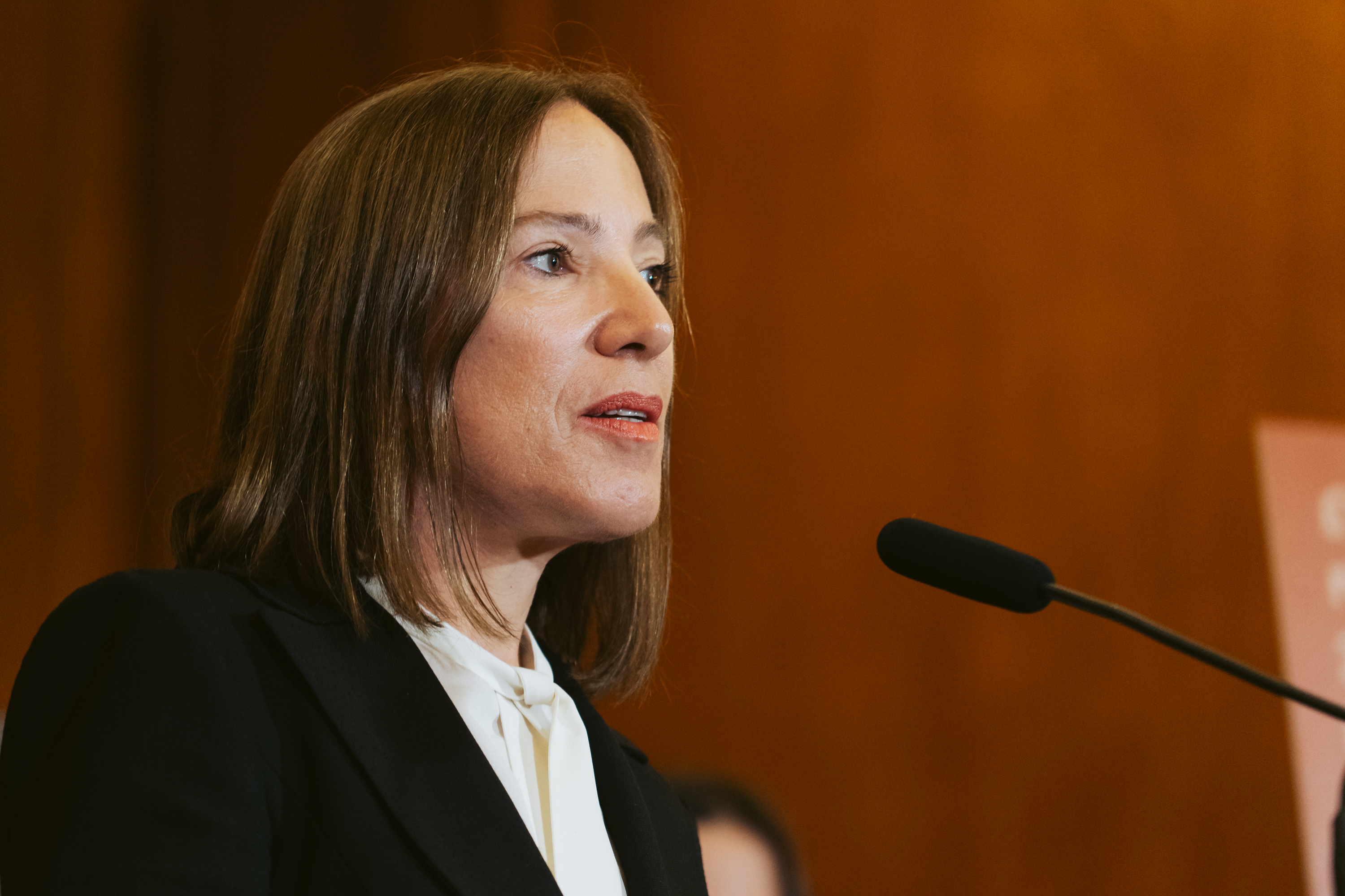 A woman with straight brown hair is speaking into a black microphone. She is wearing a white blouse and a black blazer. The background is brown with a slight blur.