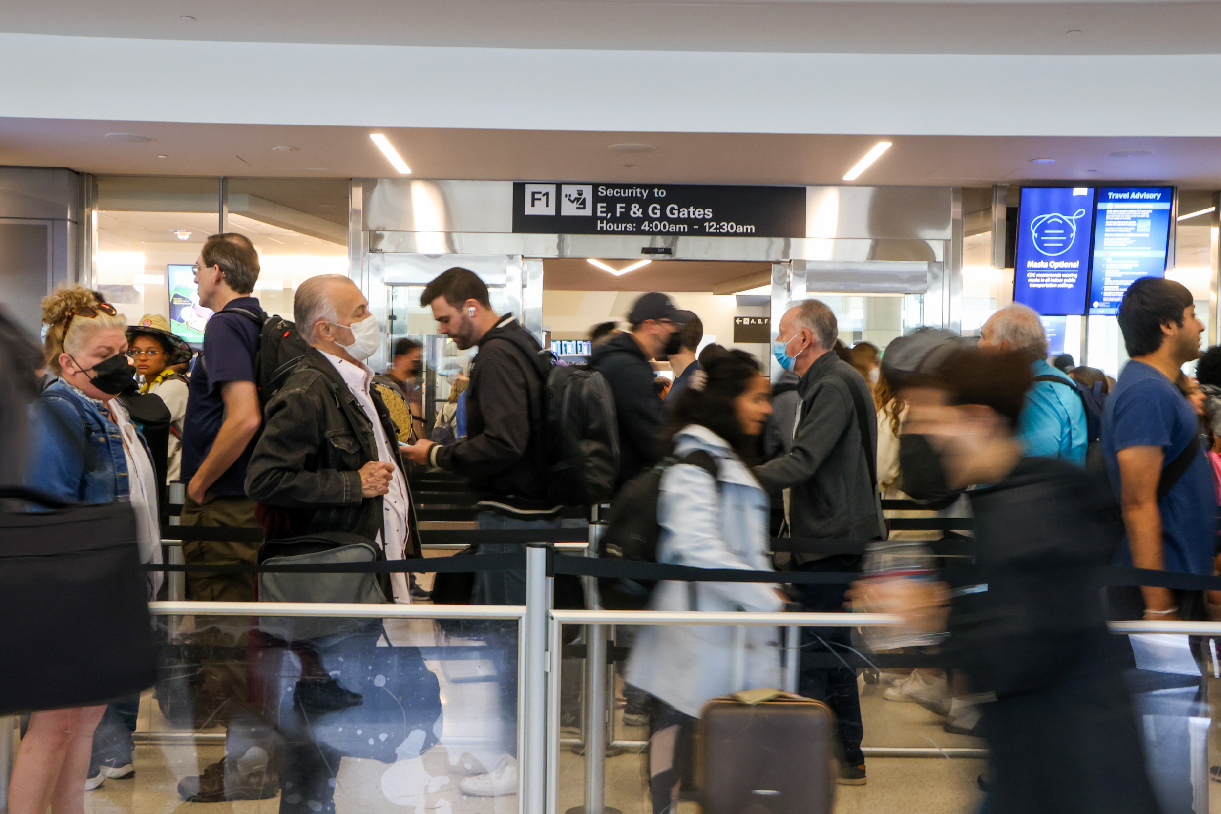 This image shows a crowded airport security checkpoint line with people wearing masks, carrying bags, and standing in queues under a sign for gates E, F, and G.