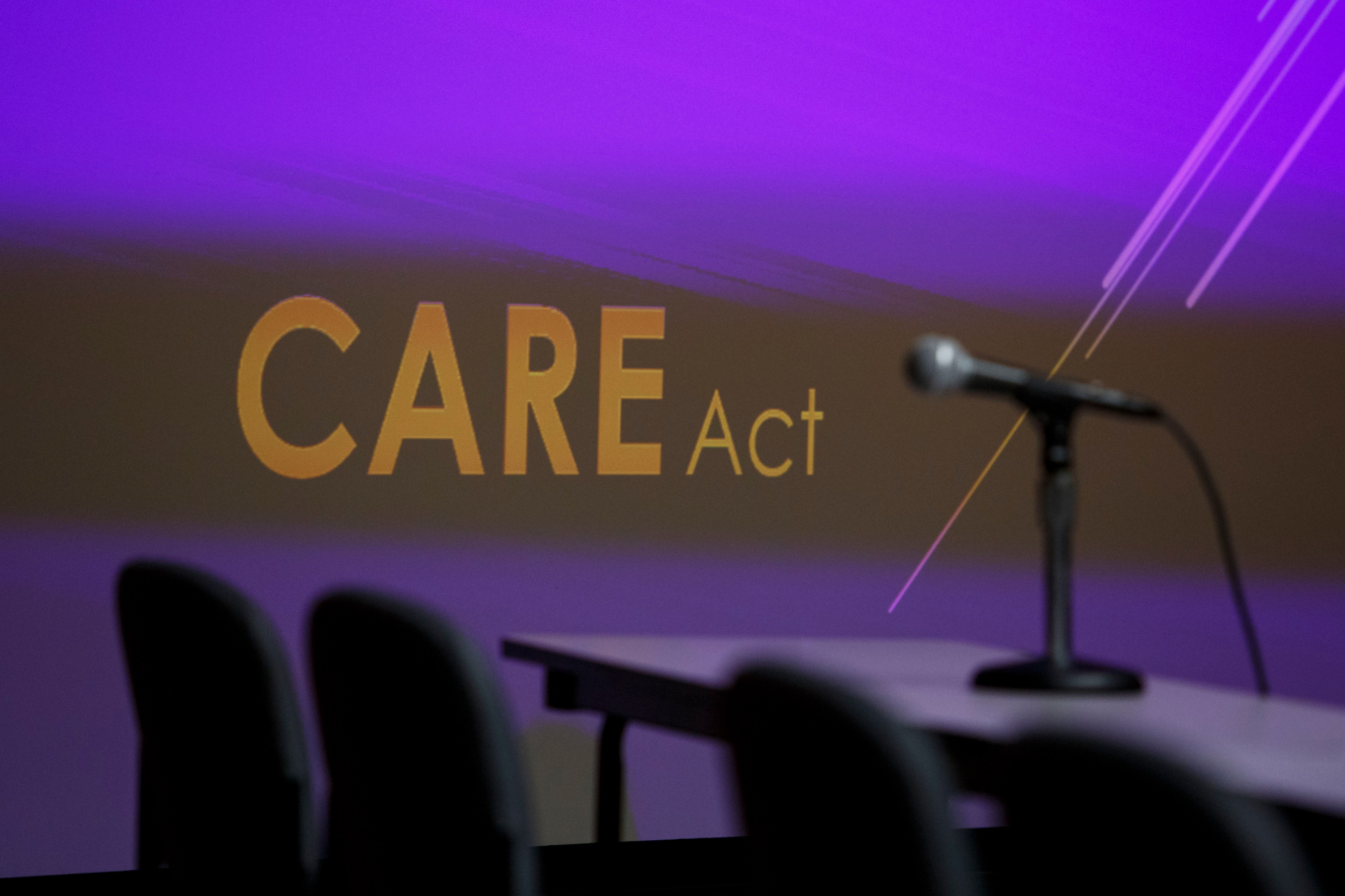 Three empty chairs and a microphone on a table seen in front of a purple backdrop that says CARE Act