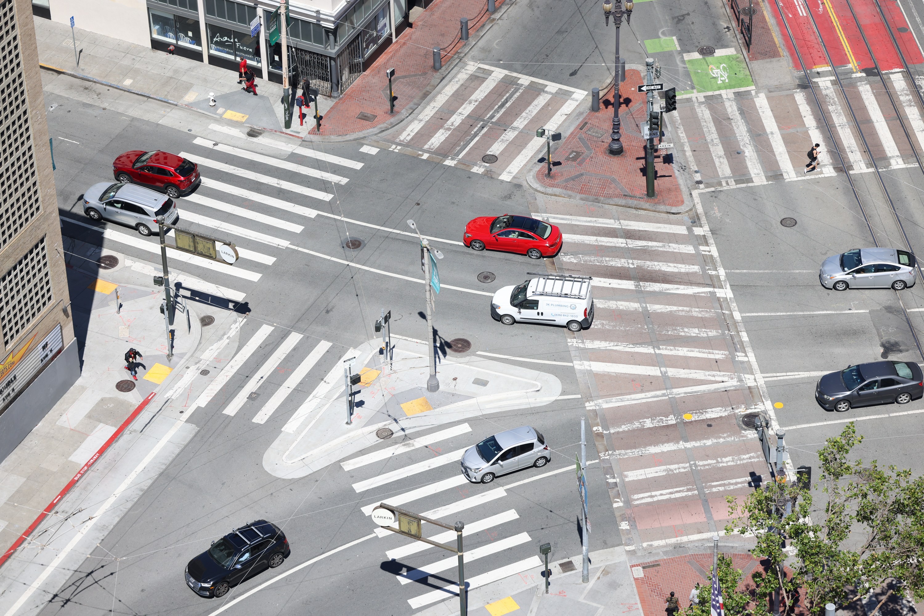 An aerial view of a busy urban intersection with multiple cars, including a red car, a white van, and several pedestrians crossing the streets in various directions.