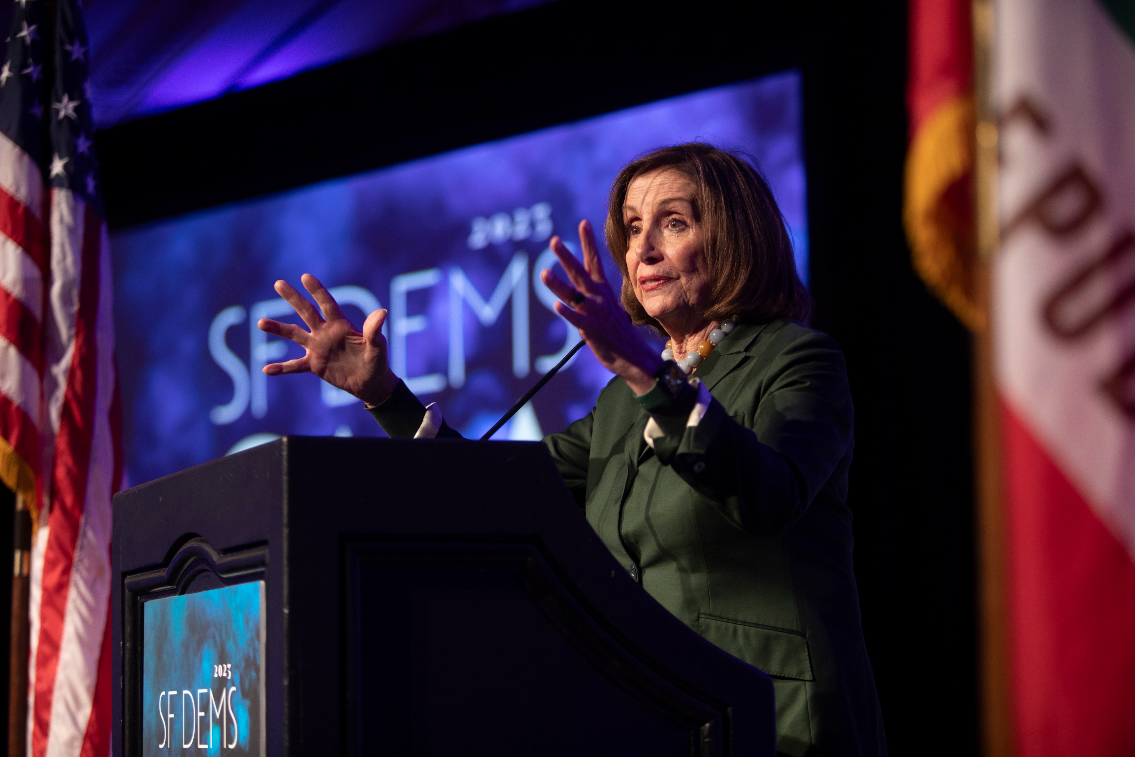 A woman in a green suit speaks passionately at a podium with &quot;2023 SF DEMS&quot; on it, flanked by the American and California flags. A purple-lit backdrop is behind her.