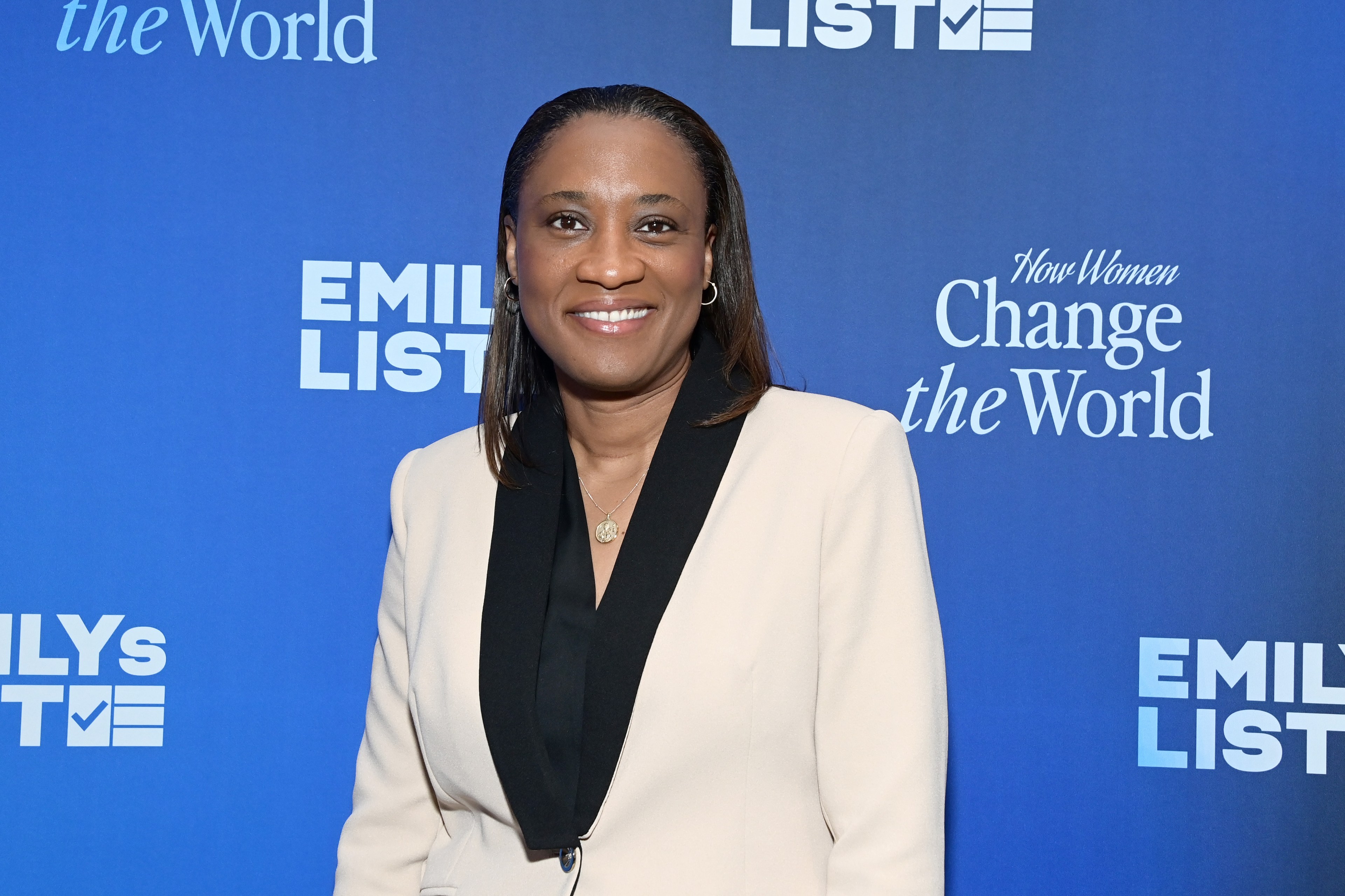 A smiling woman in a beige blazer stands before a blue backdrop with the text "EMILY's LIST" and "How Women Change the World."