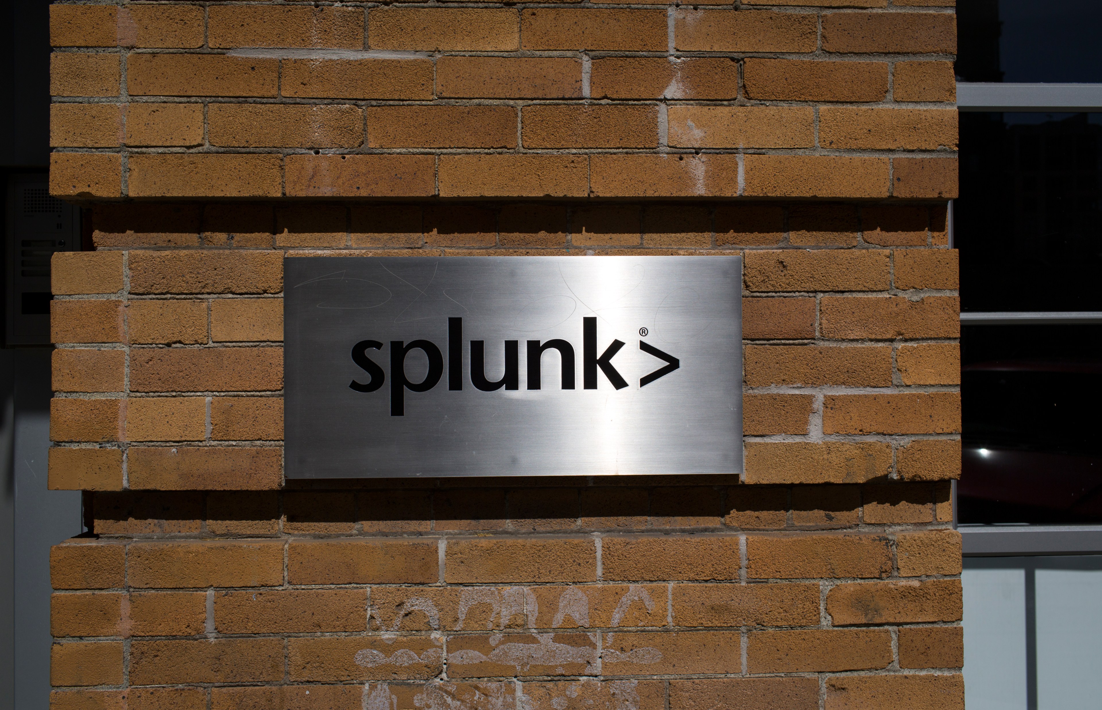 A silver-colored brushed metal sign mounted to a brick wall shines in sunlight. On the sign in lower case black lettering is a company logo.