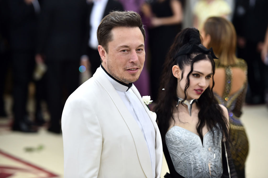 Elon Musk, wearing a white suit and Grimes, wearing a silver dress, are seen from the side standing close together in a photo showing them attending the Heavenly Bodies: Fashion and the Catholic Imagination Costume Institute Gala at The Metropolitan Museum of Art on May 7, 2018, in New York City.