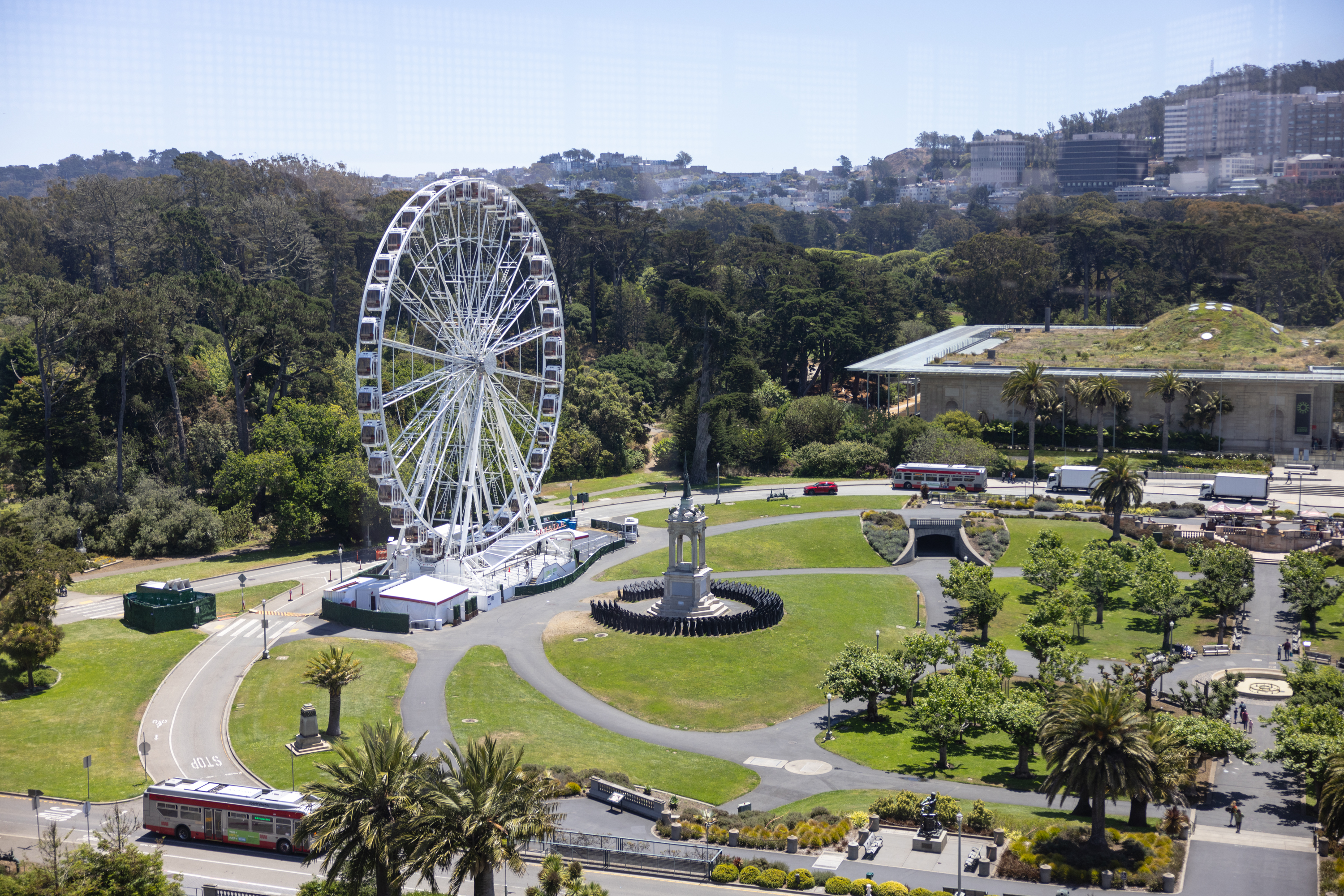 A Ferris wheel in a park with lush trees, paths, a sculpture, and a tram passing by.