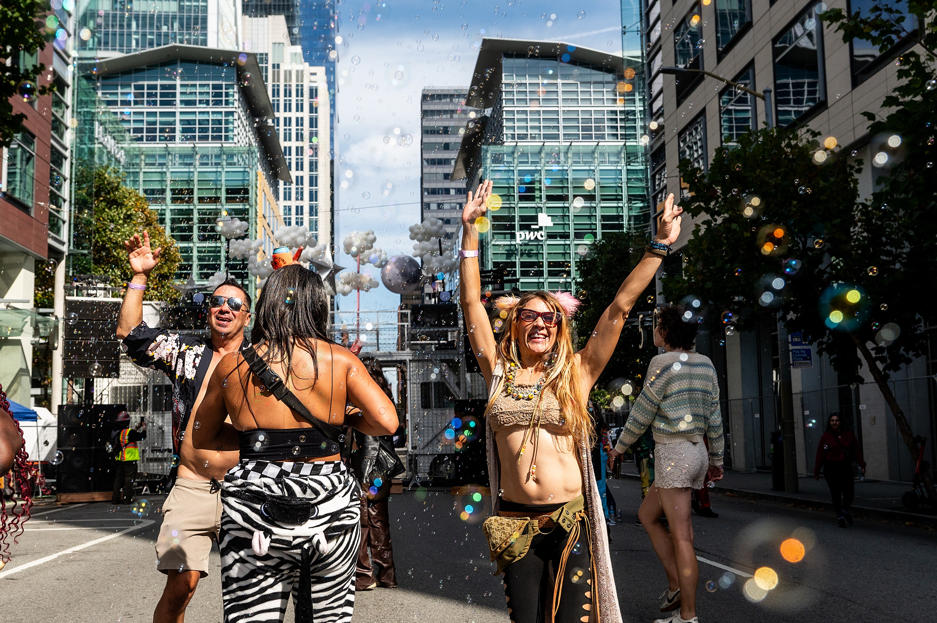 People joyfully dance on a city street surrounded by tall buildings. They wear colorful, festive outfits, and bubbles float around them in the sunlight.