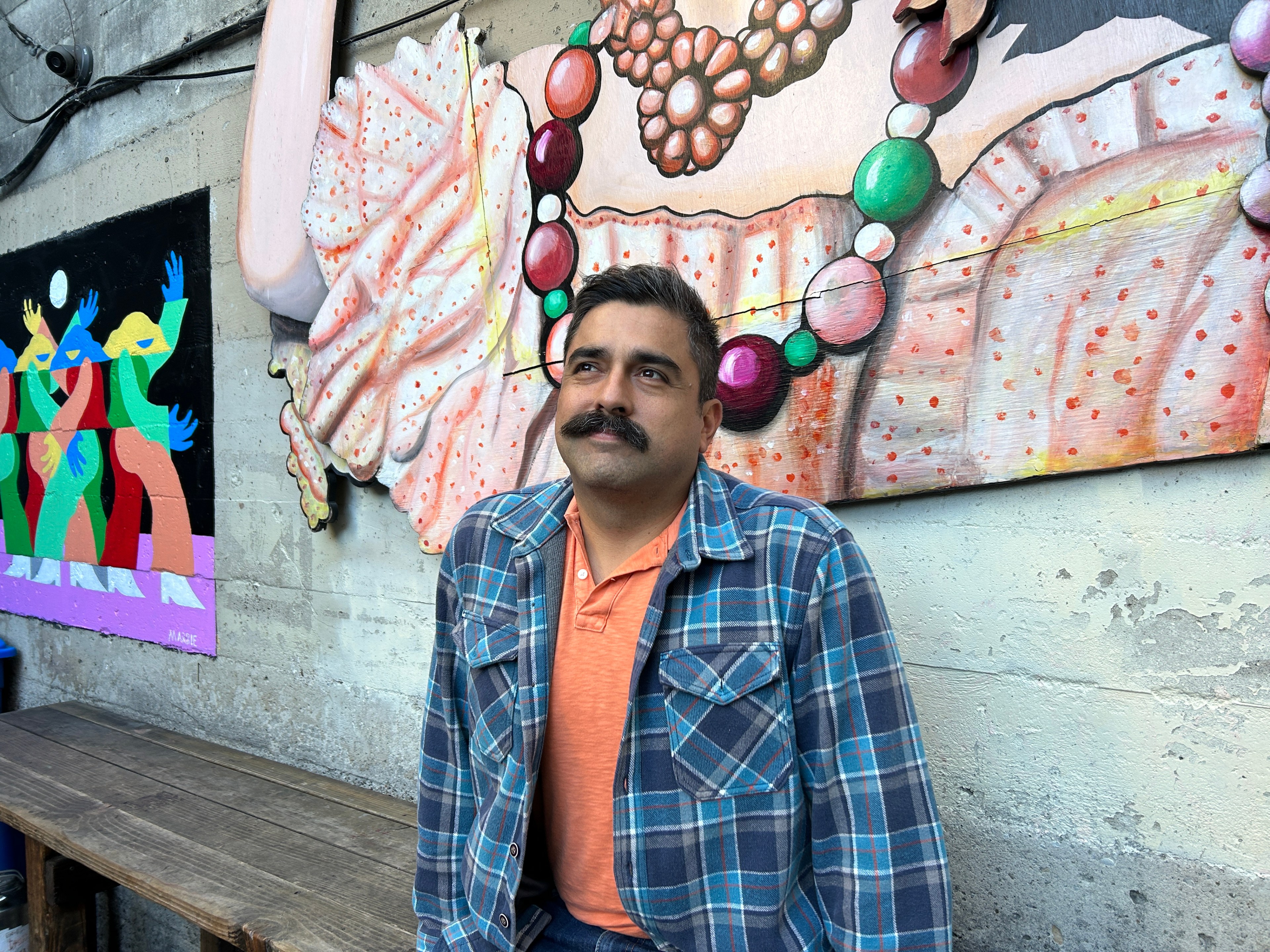 A man sits before a colorful mural with abstract festive elements and a painting of dancing figures.