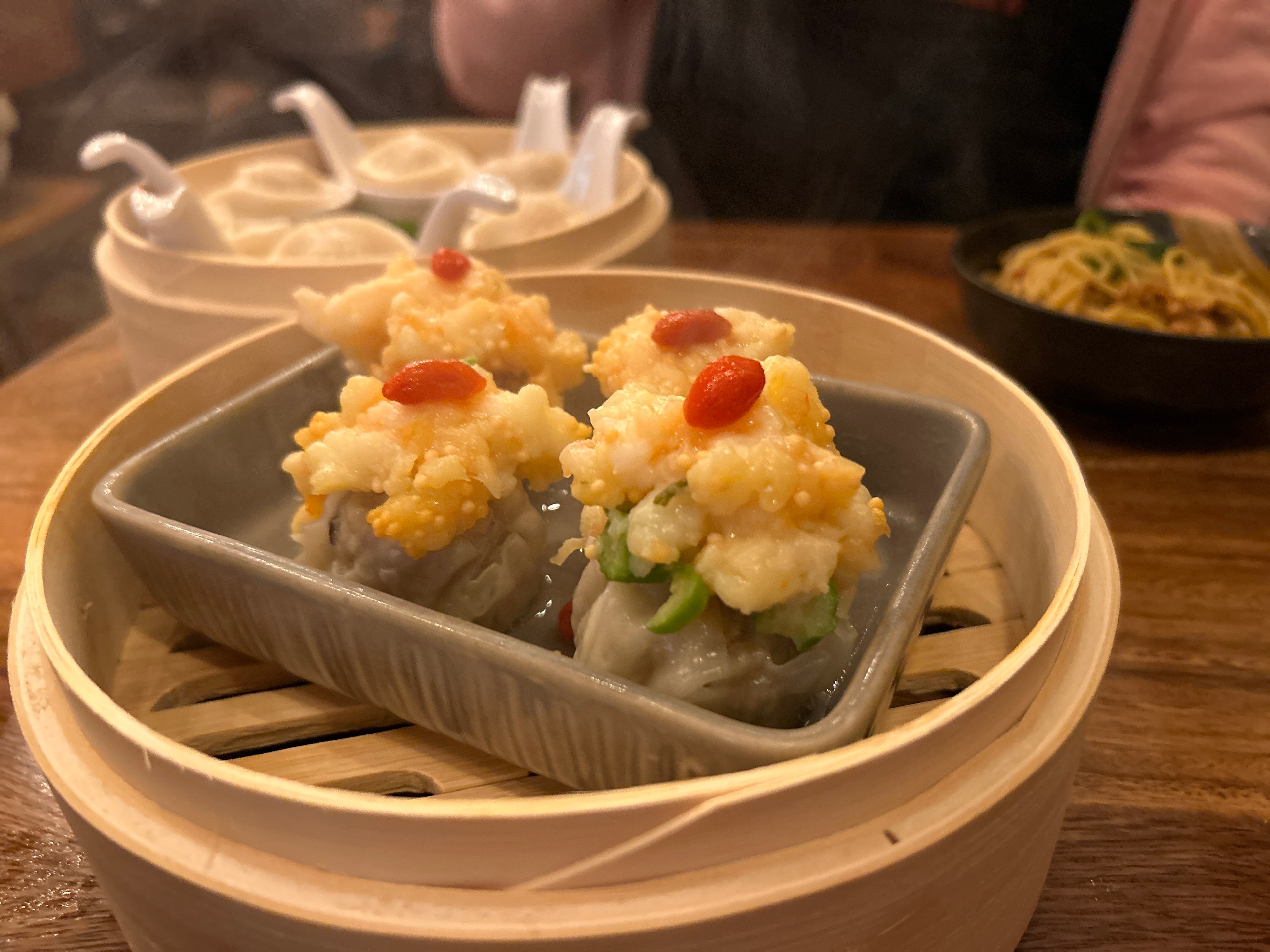 Pork shumai sits in a dim sum basket on a table with other East Asian dishes.