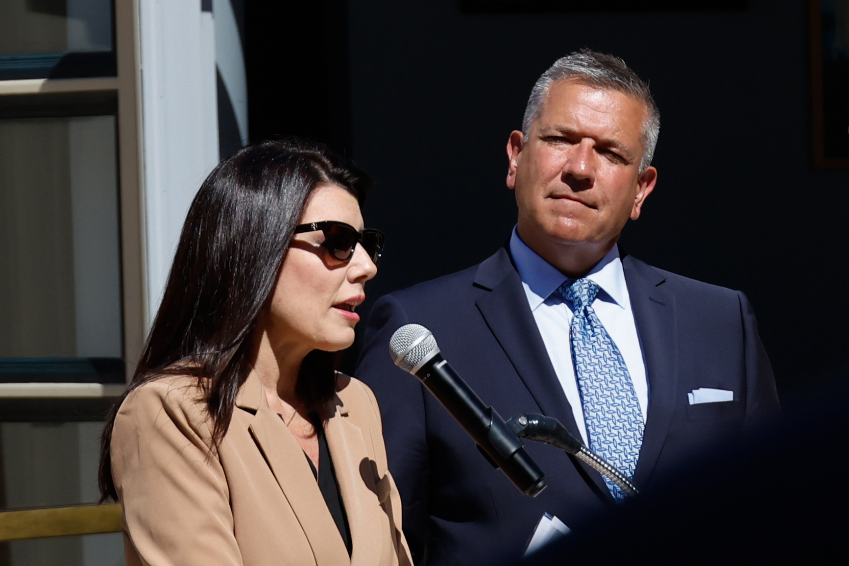 A man and woman, both in professional attire, stand beside a microphone under sunlight.
