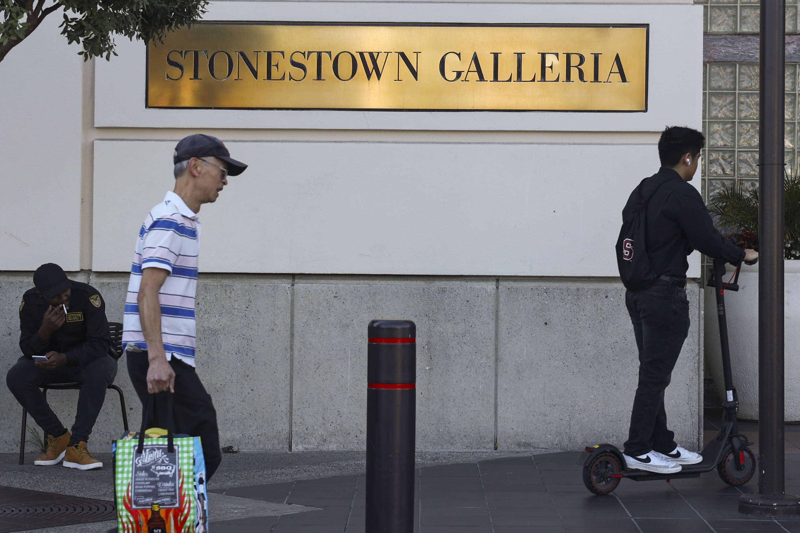 Two shoppers walk past a brass plaque that reads, "Stonestown Galleria."