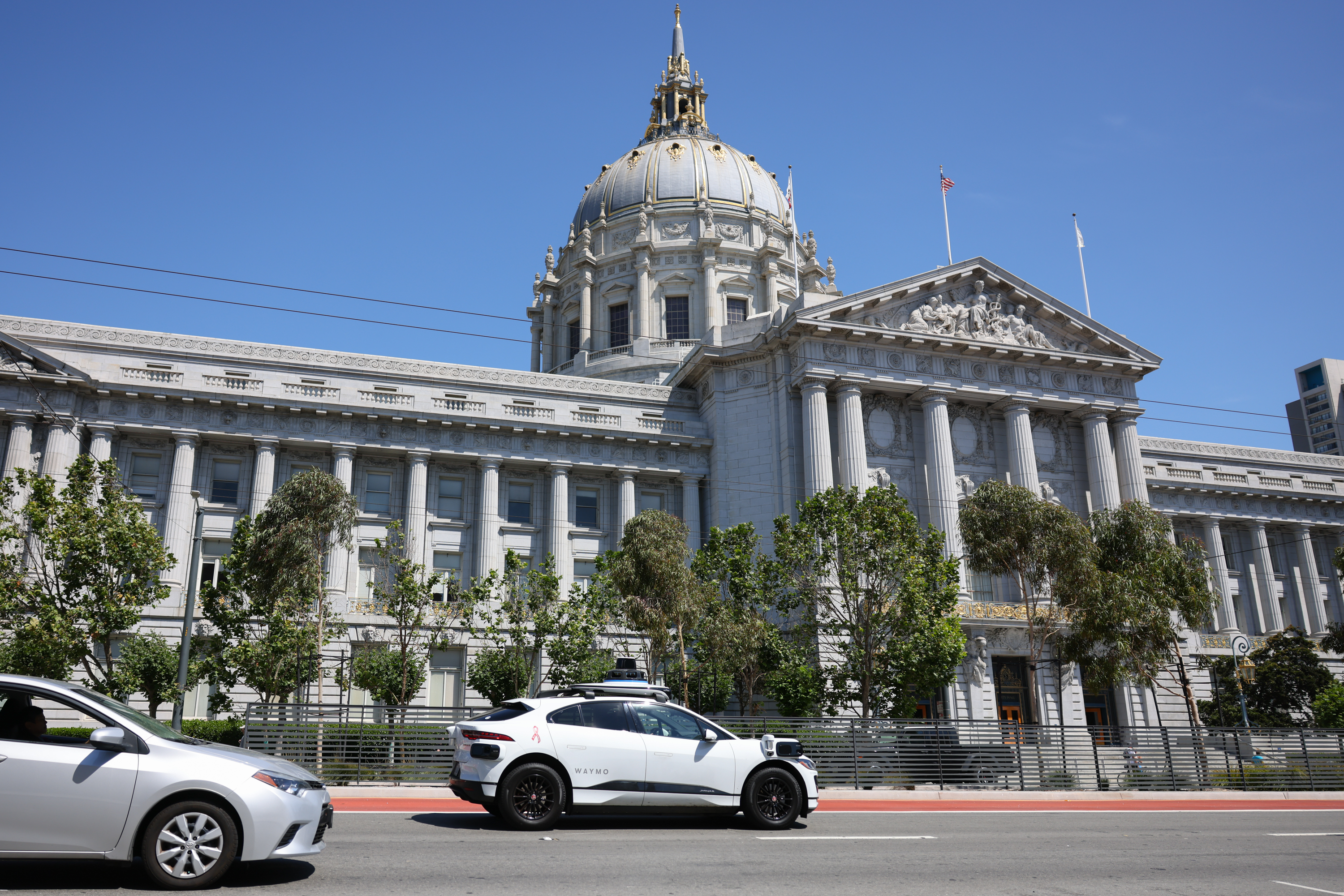 A Waymo is on the road in front of San Francisco City Hall. Another gray sedan is also visible on the left-bottom side of the photo