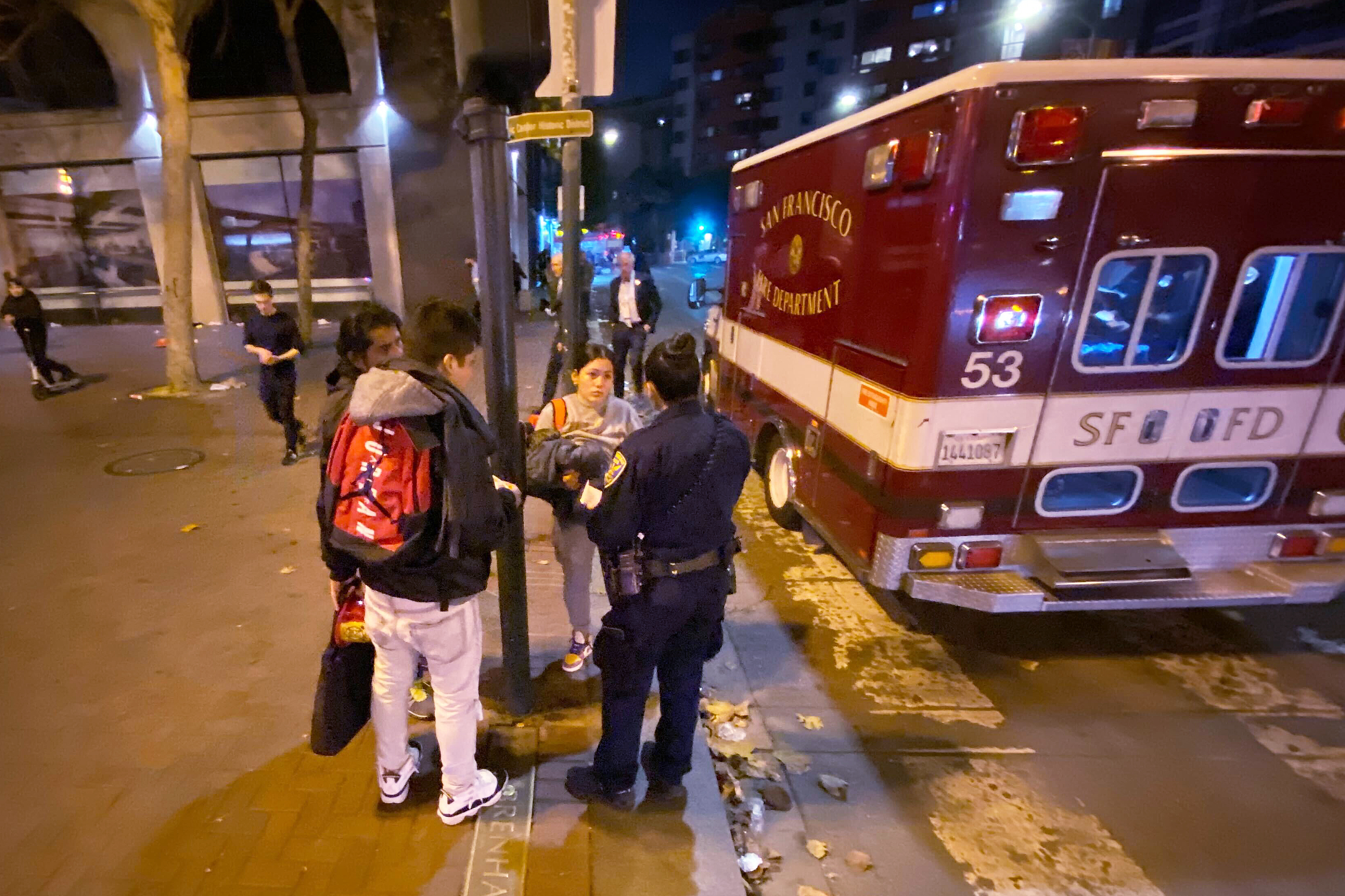 An ambulance is parked on a city street at night, with a police officer talking to two people next to it.