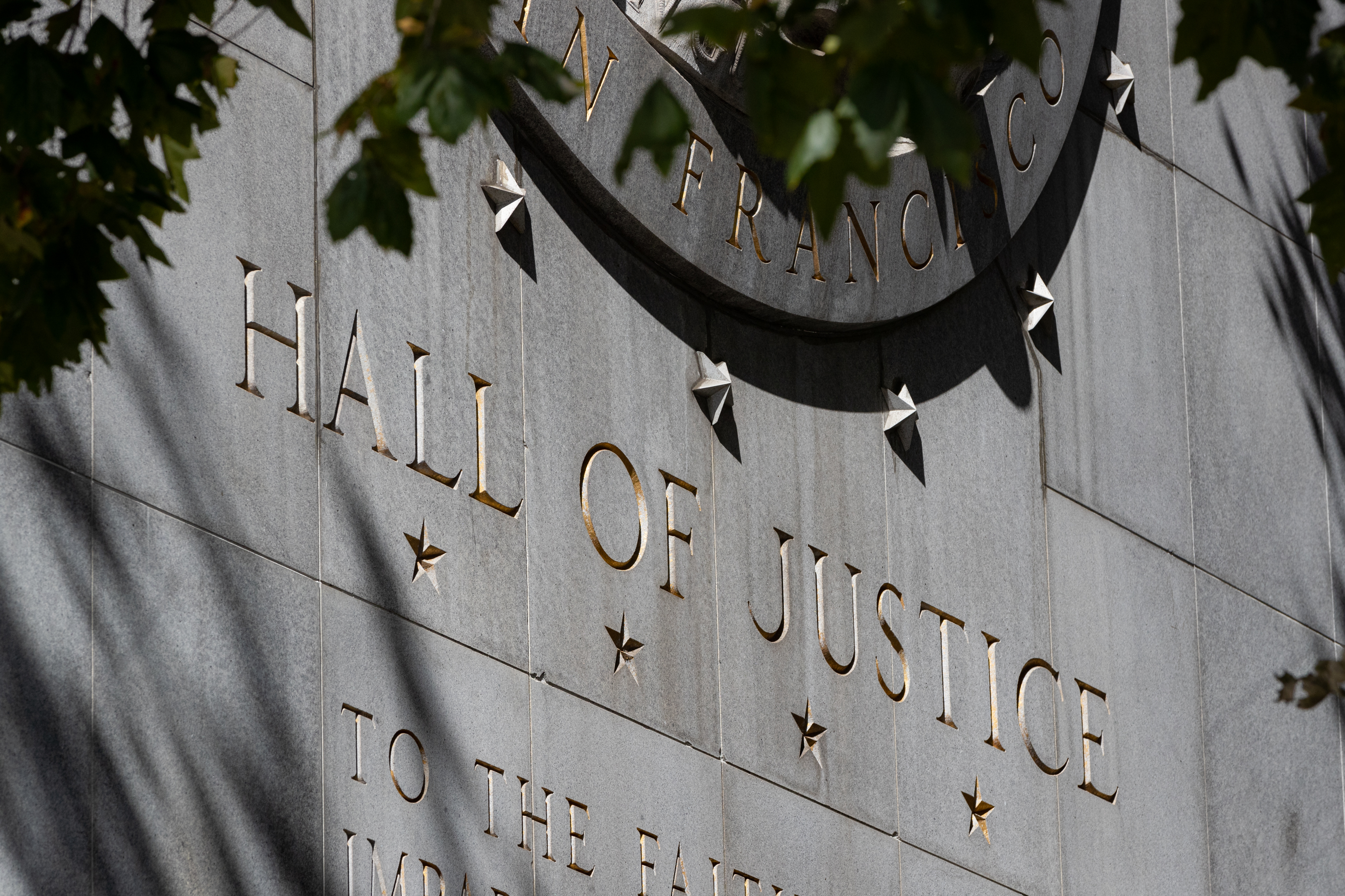 A sunlit stone wall with "HALL OF JUSTICE" engraved, adorned with stars, shadow of a tree's leaves visible.