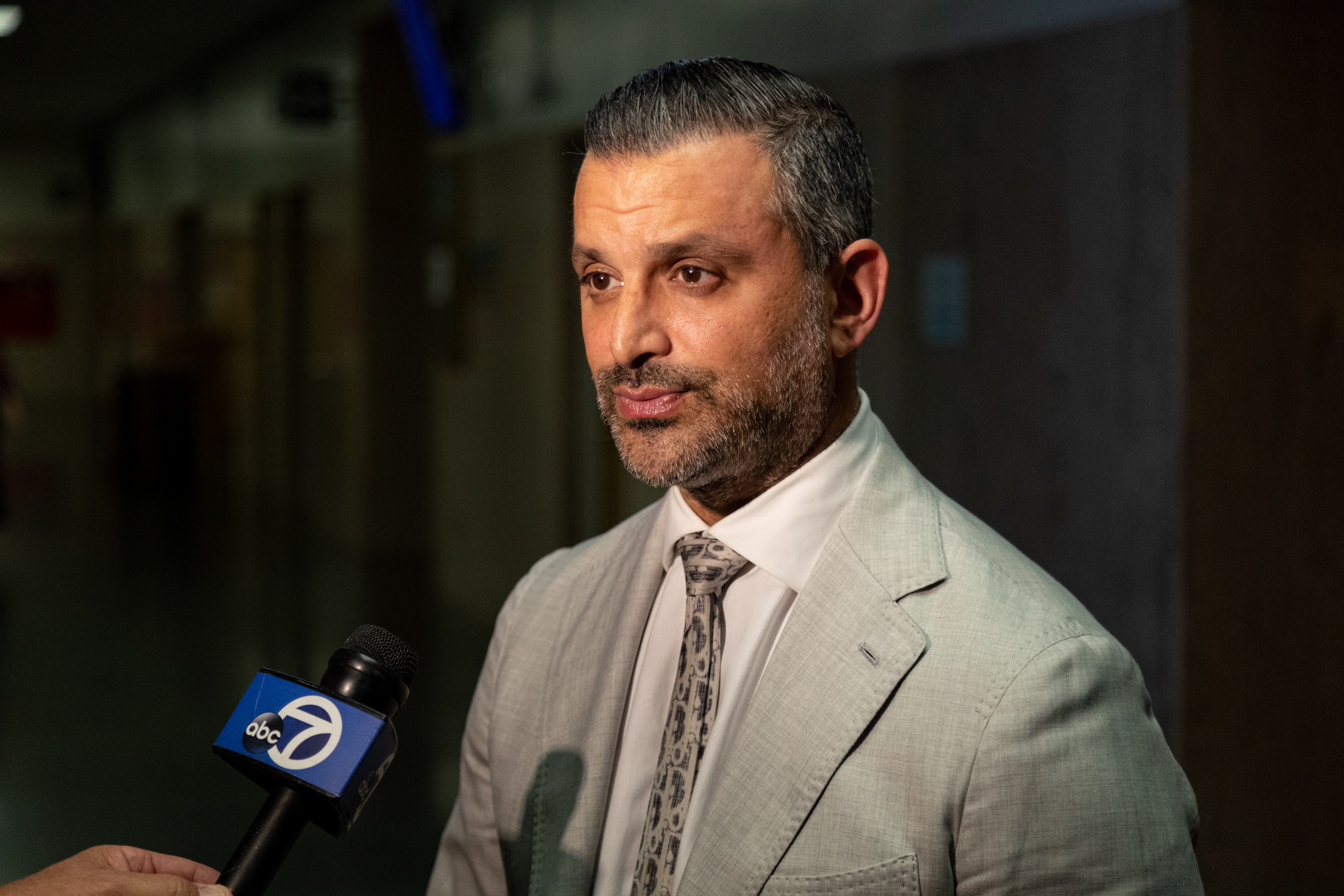 A man in a light suit and tie is speaking in a dim hallway. A microphone marked &quot;abc 7&quot; is held towards him.