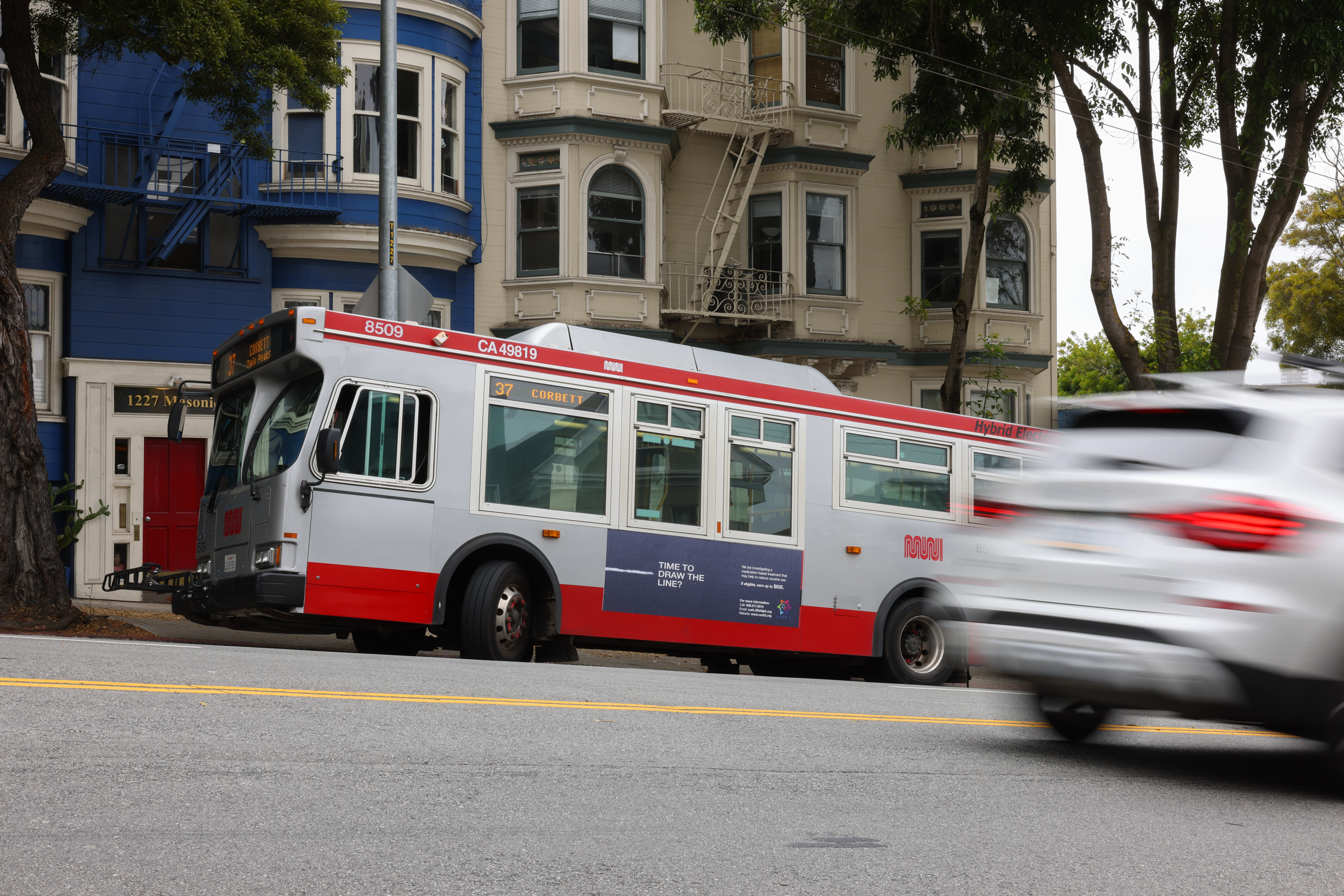 A city bus parked by the curb with a blurred car passing by in front, and classic houses in the background.