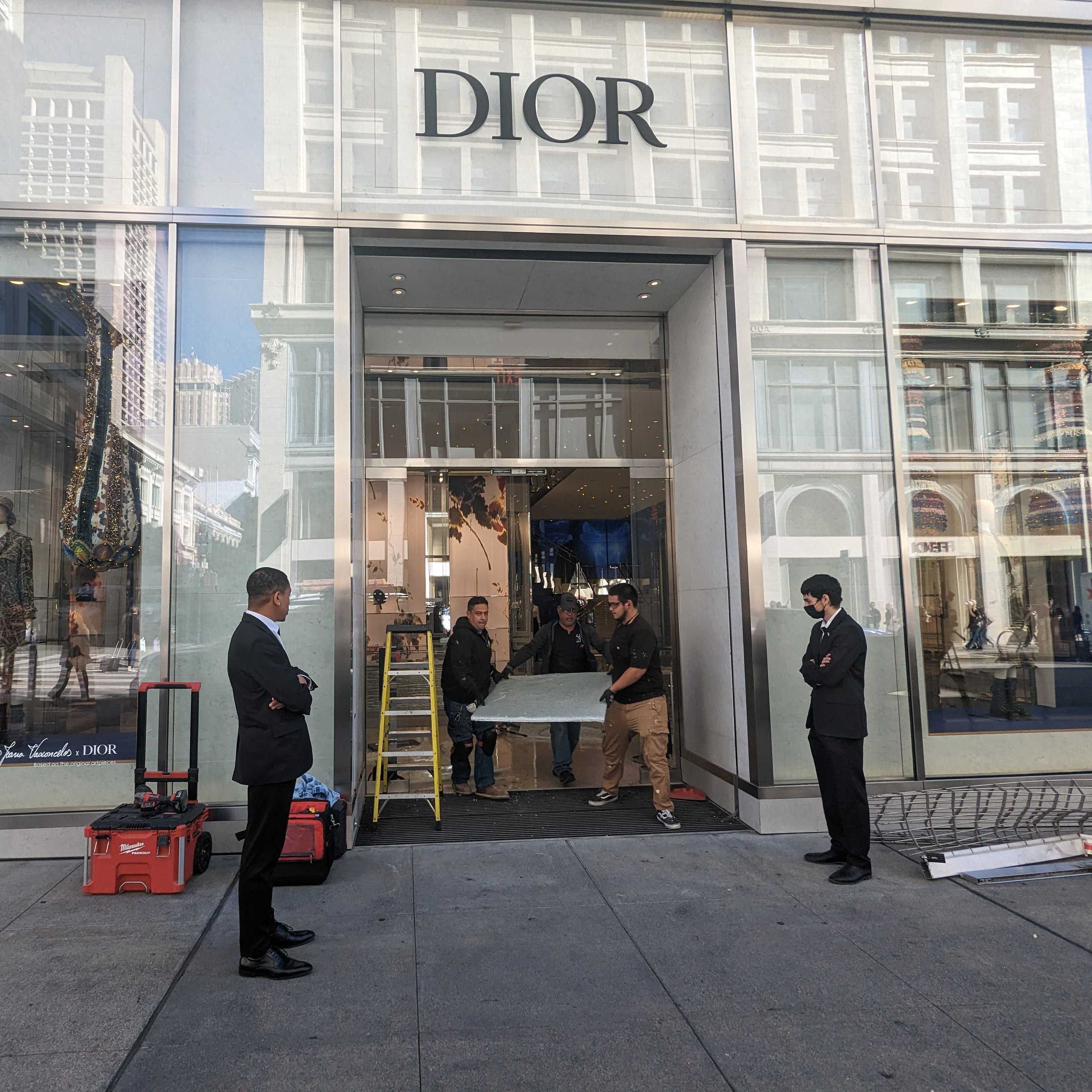 Security guards stand near the entrance to a luxury clothing store while workmen fix the doorway.