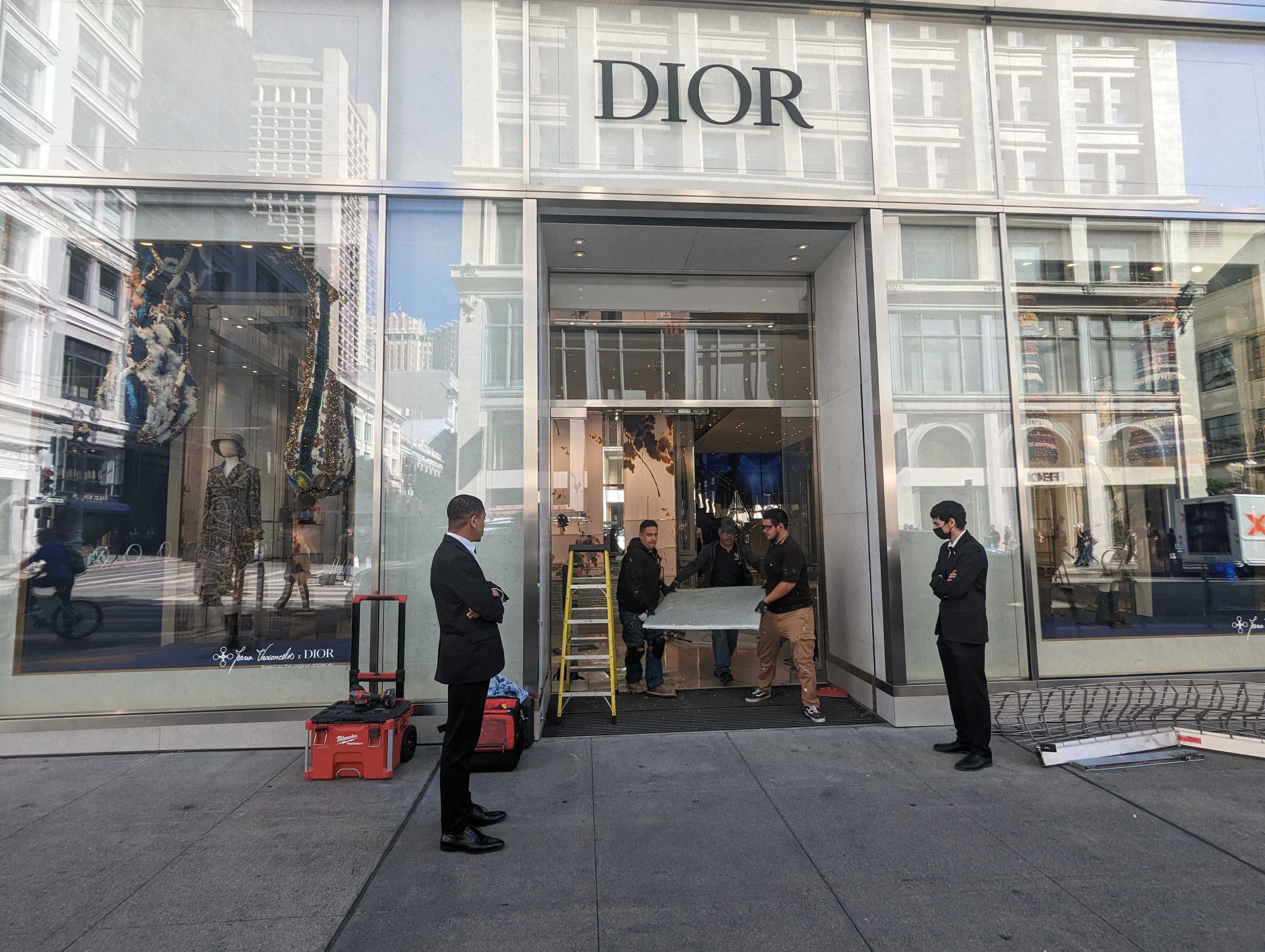 Security guards stand near the entrance to a luxury clothing store while workmen fix the doorway.