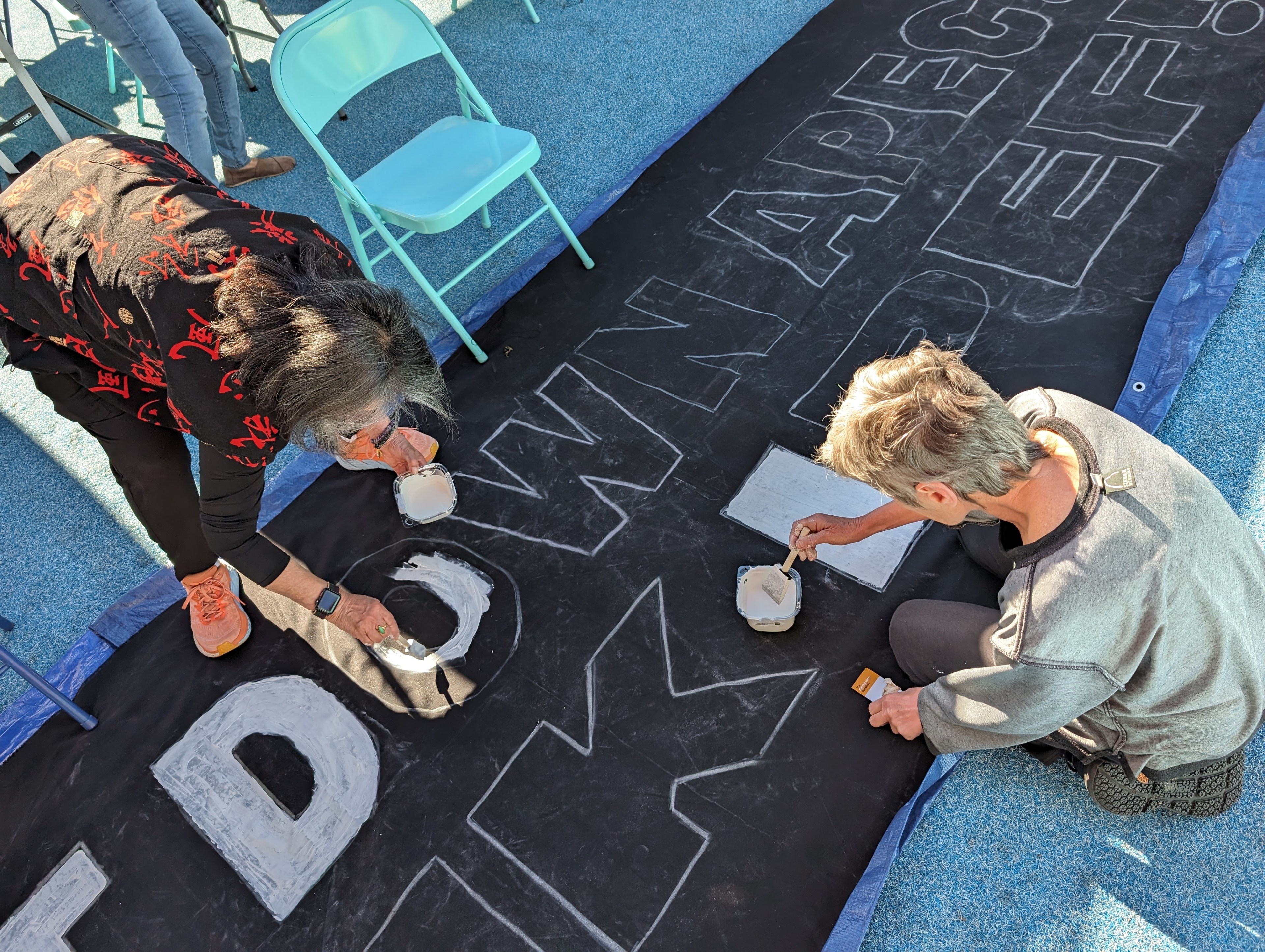 Pam Tau Lee and Kathe Burick paint letters on a "Shut down APEC" sign Sunday at a teach-in at Kawpa Gardens on Mission Street.