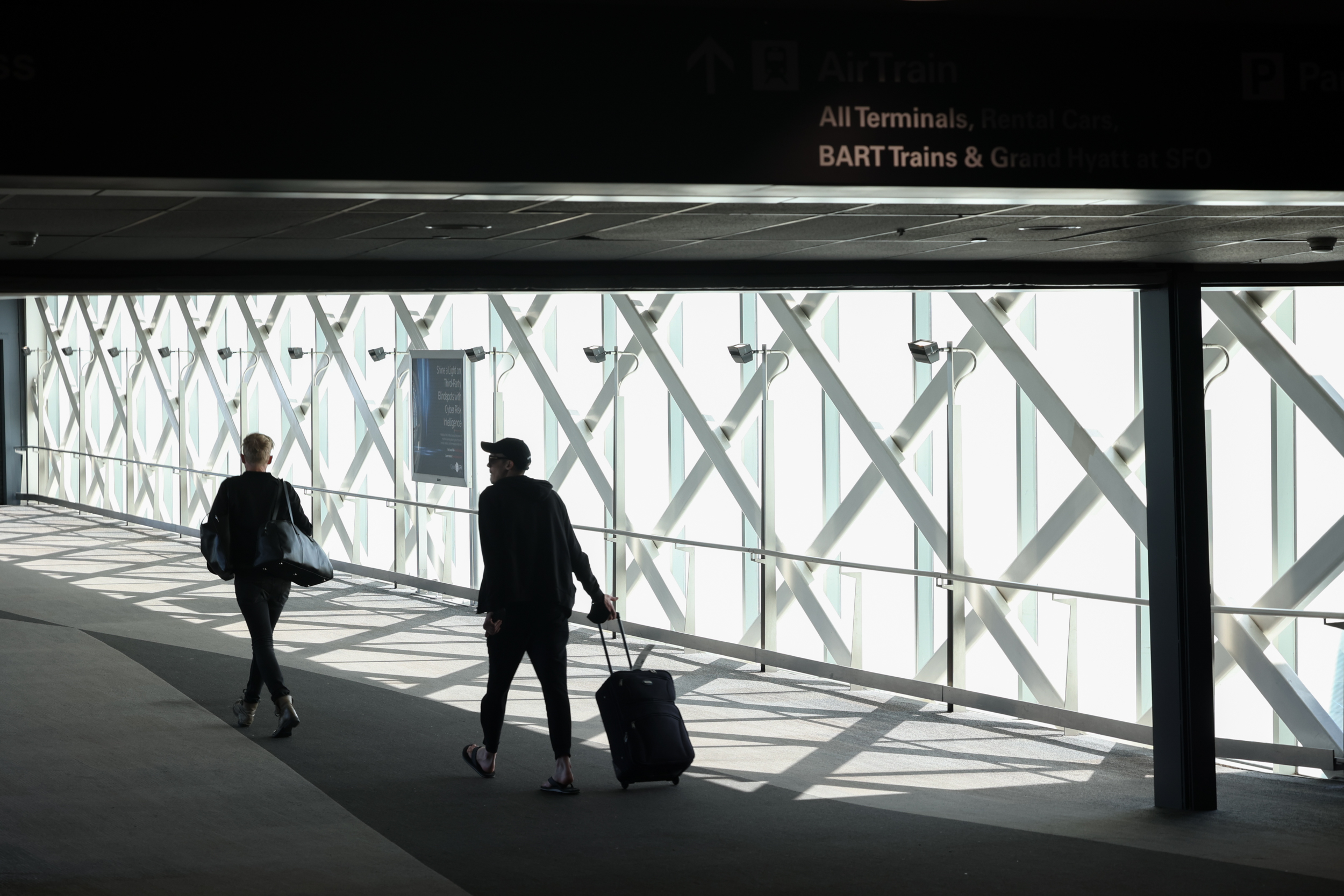 Two travelers, silhouetted against strong exterior light, walk along the gray carpeted bridgeway leading into San Francisco International. Airport.