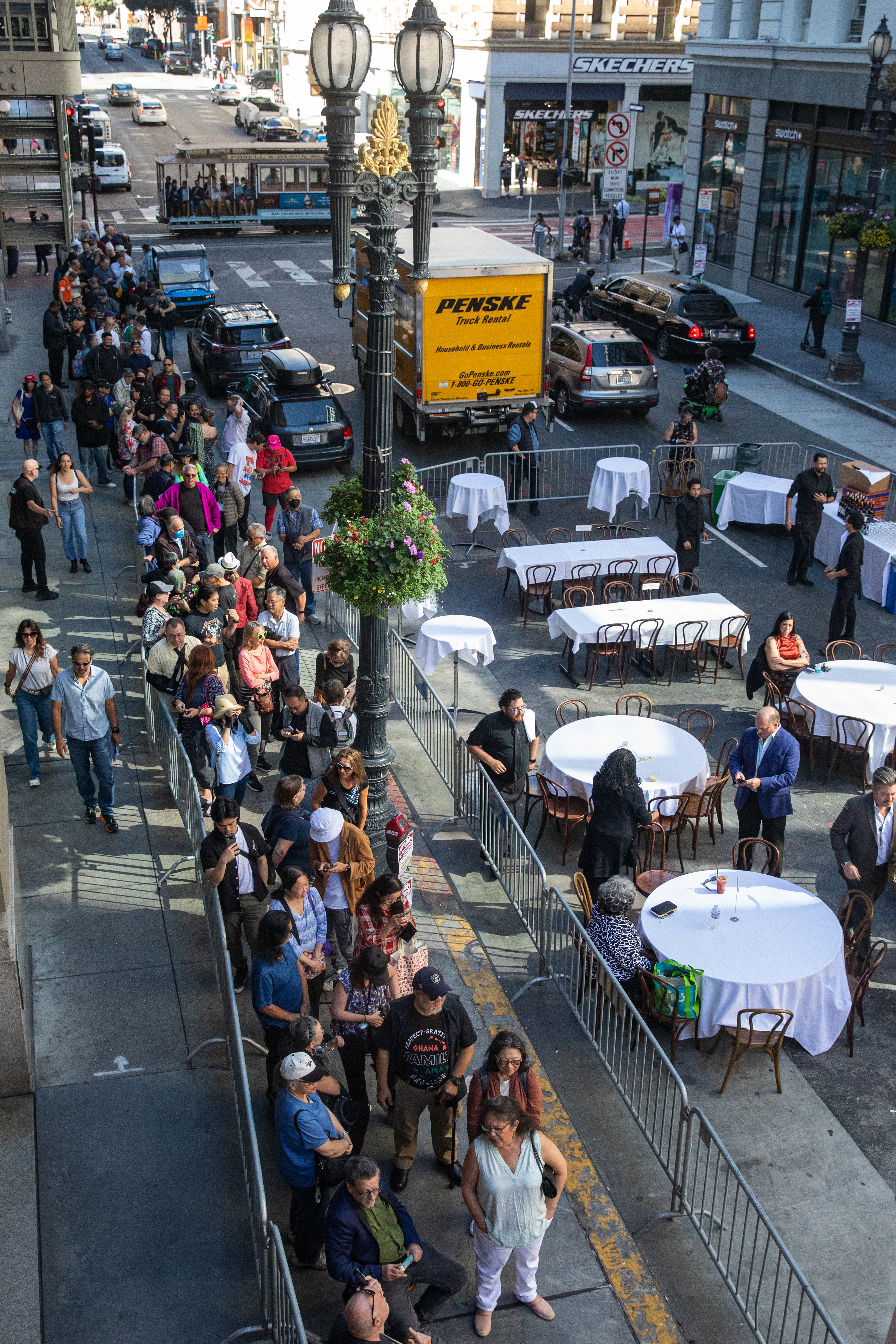 A large crowd is lined up on a city street beside a dining area with tables covered in white tablecloths. A yellow truck and cable car are in the background.