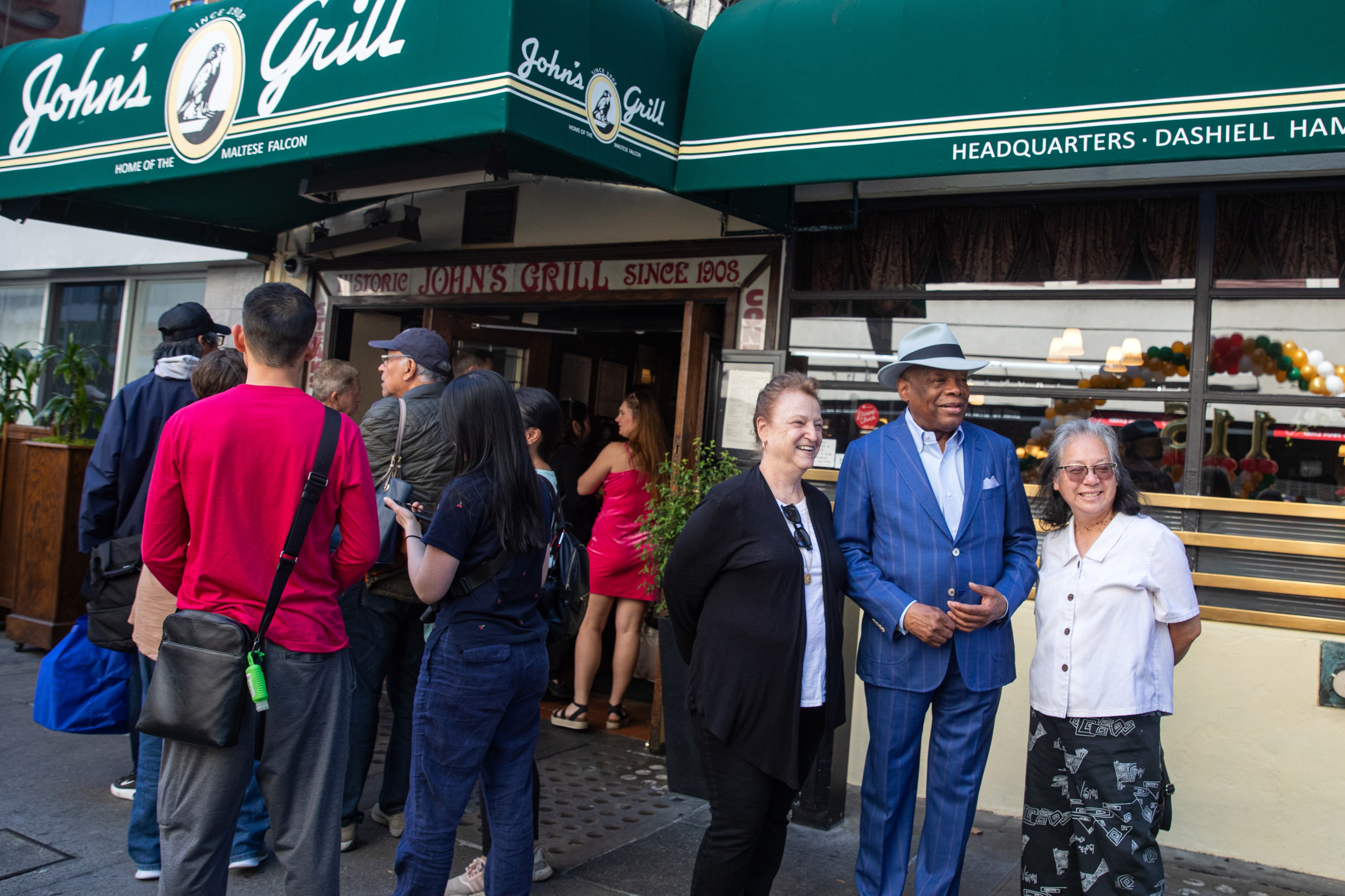 A group of people wait outside John's Grill. On the right, two women and a man in a blue suit and hat stand smiling, posing for a photo.