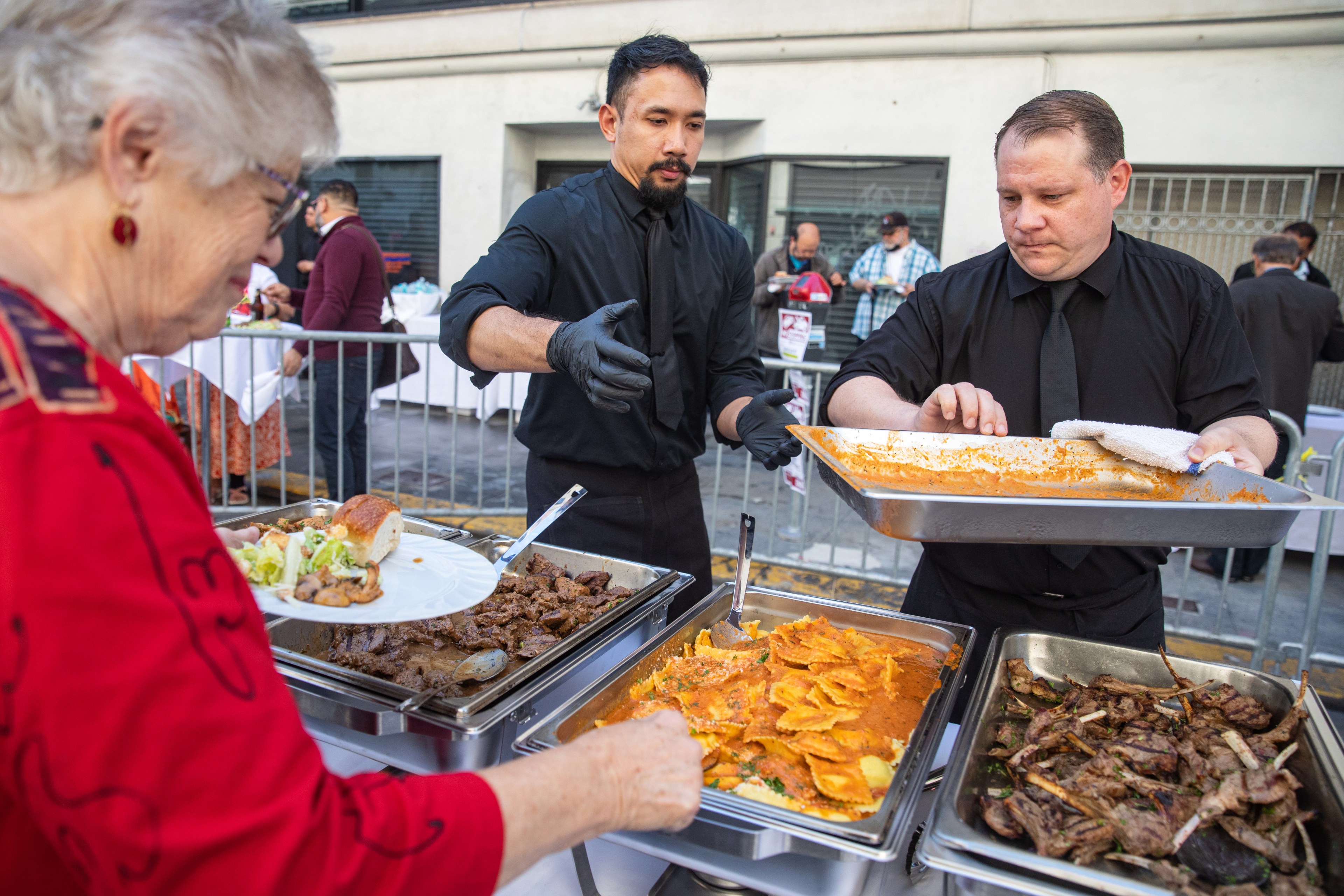 Two servers in black attire offer various dishes from chafing trays to a woman holding a plate. The scene is outdoors with other people in the background.