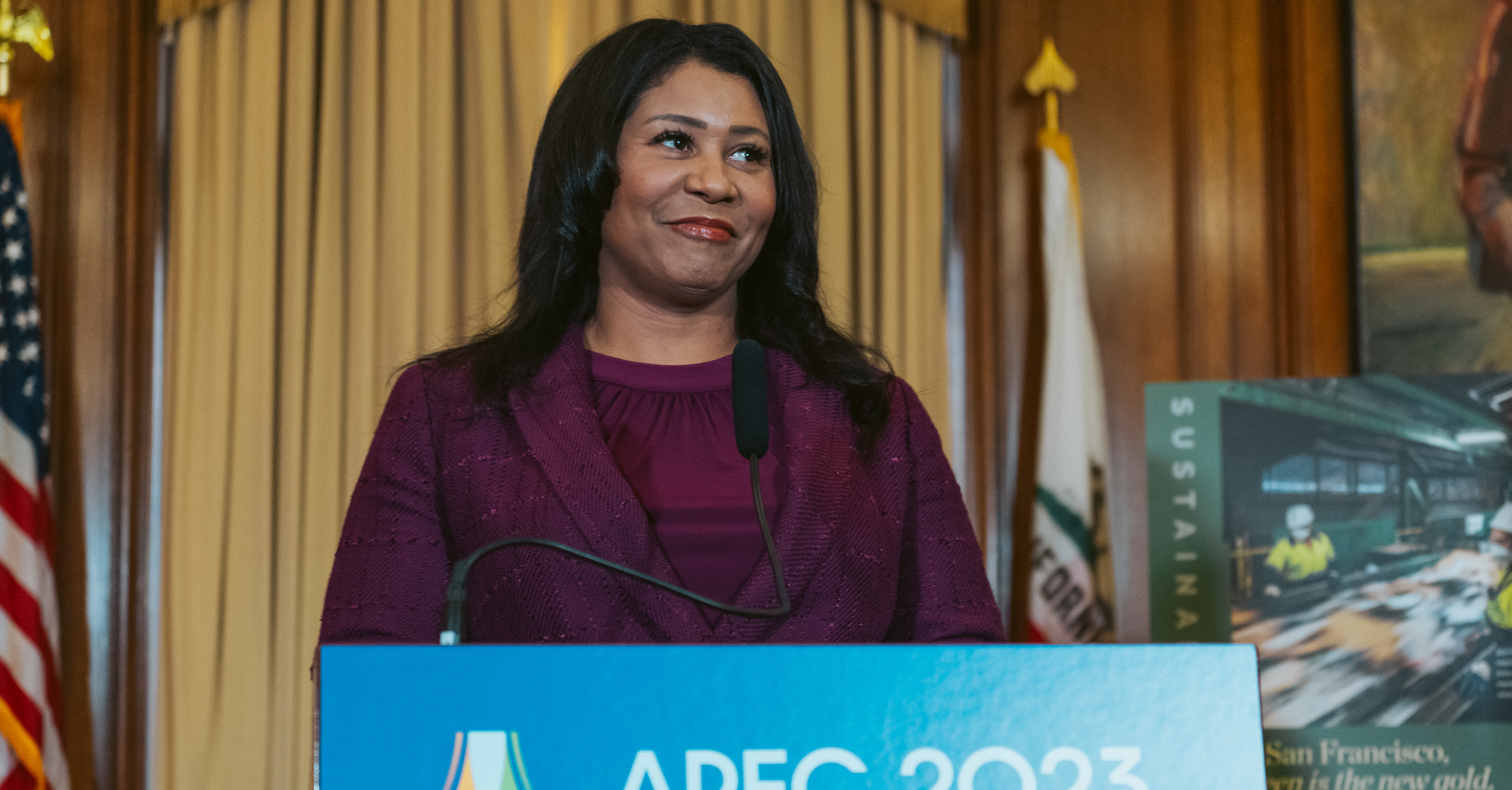 San Francisco Mayor London Breed, dressed in a purple speaks to the media during a press conference in City Hall. She stands behind a podium that reads "APEC 2023 UNITED STATES" in a wood paneled room.