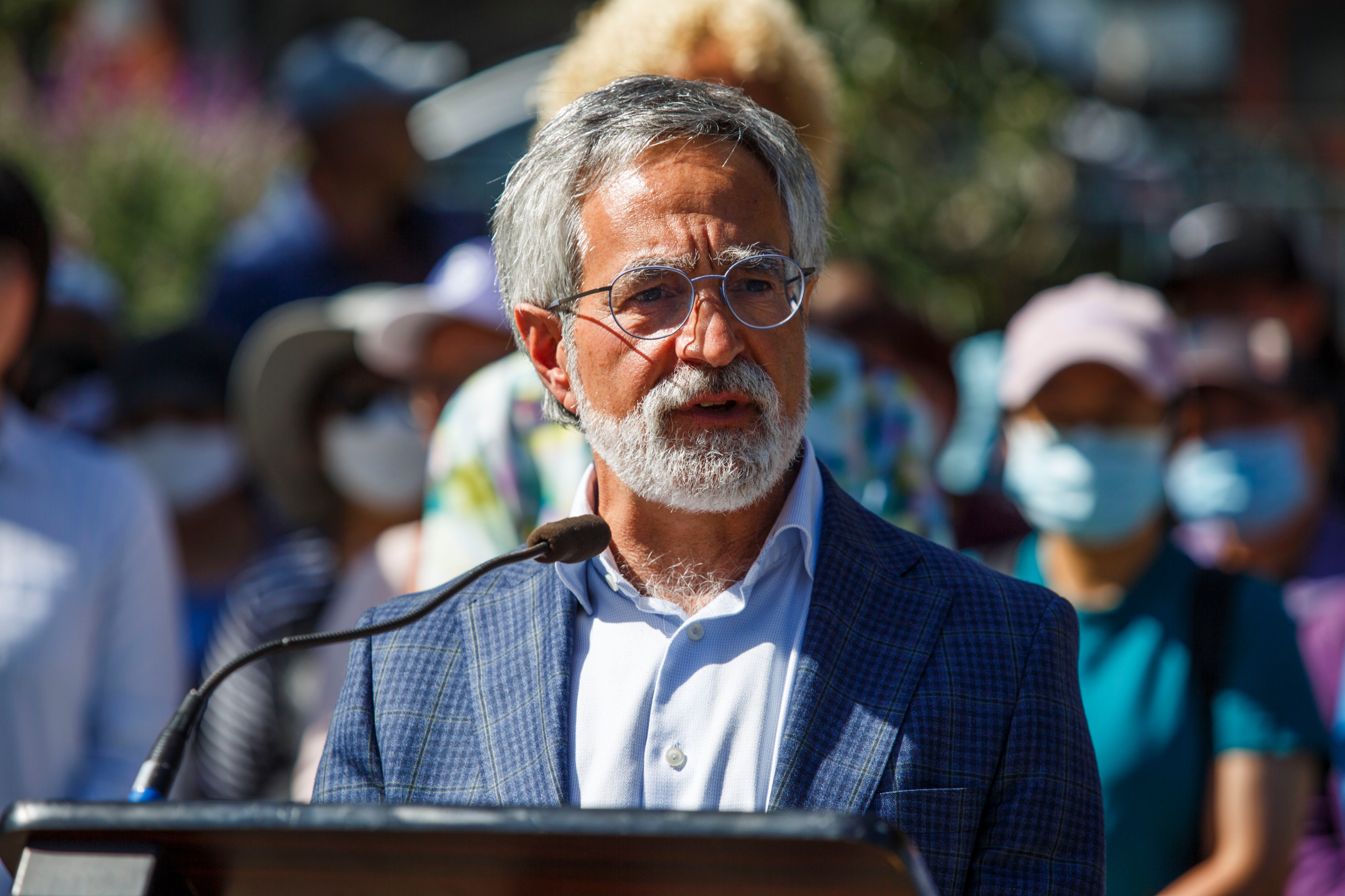 A man with gray hair and beard speaks at a podium outdoors, a crowd with masks in the background.