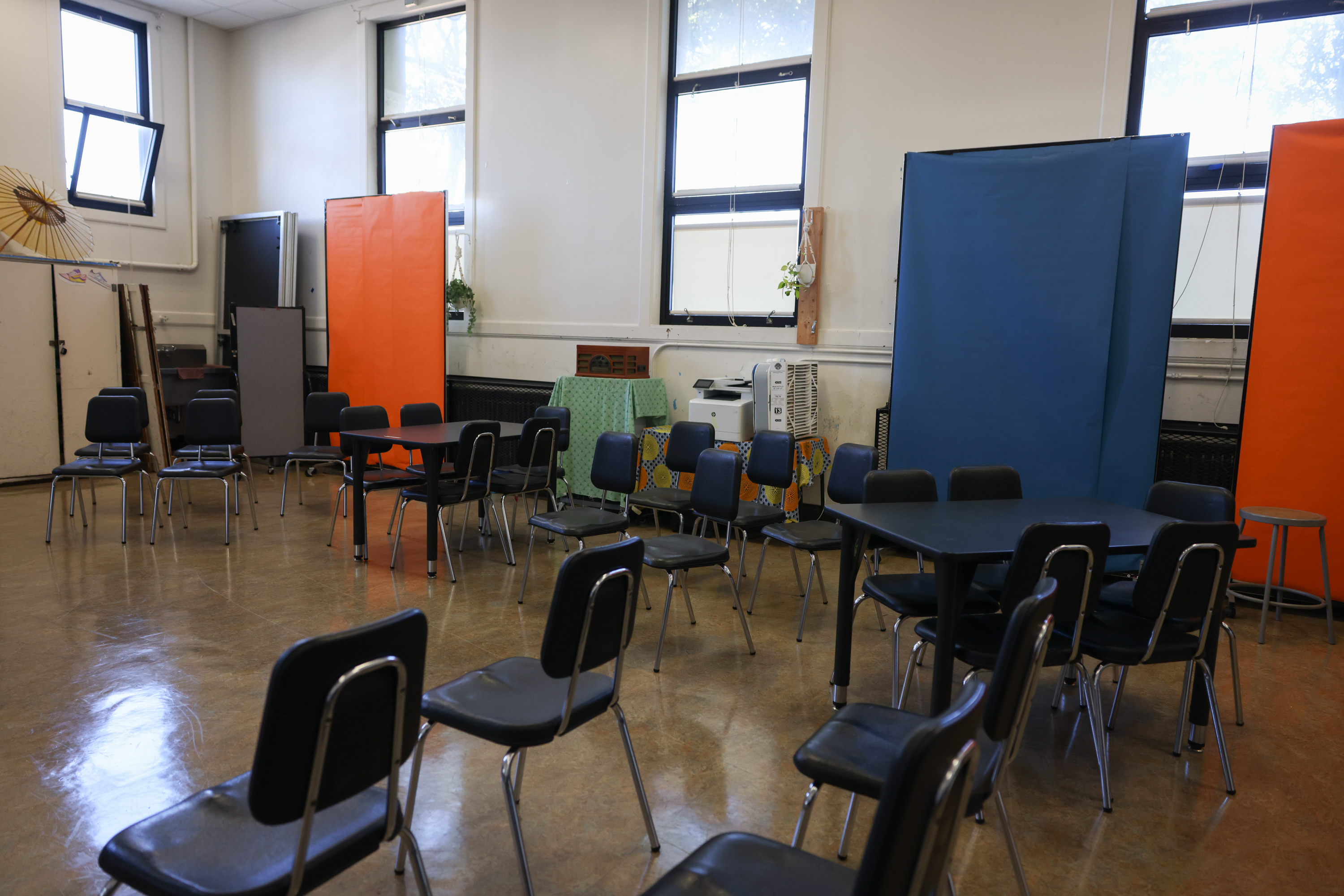 A room with scattered black chairs and tables, blue and orange partitions, three windows, plants, and a fabric-covered table with a fan.