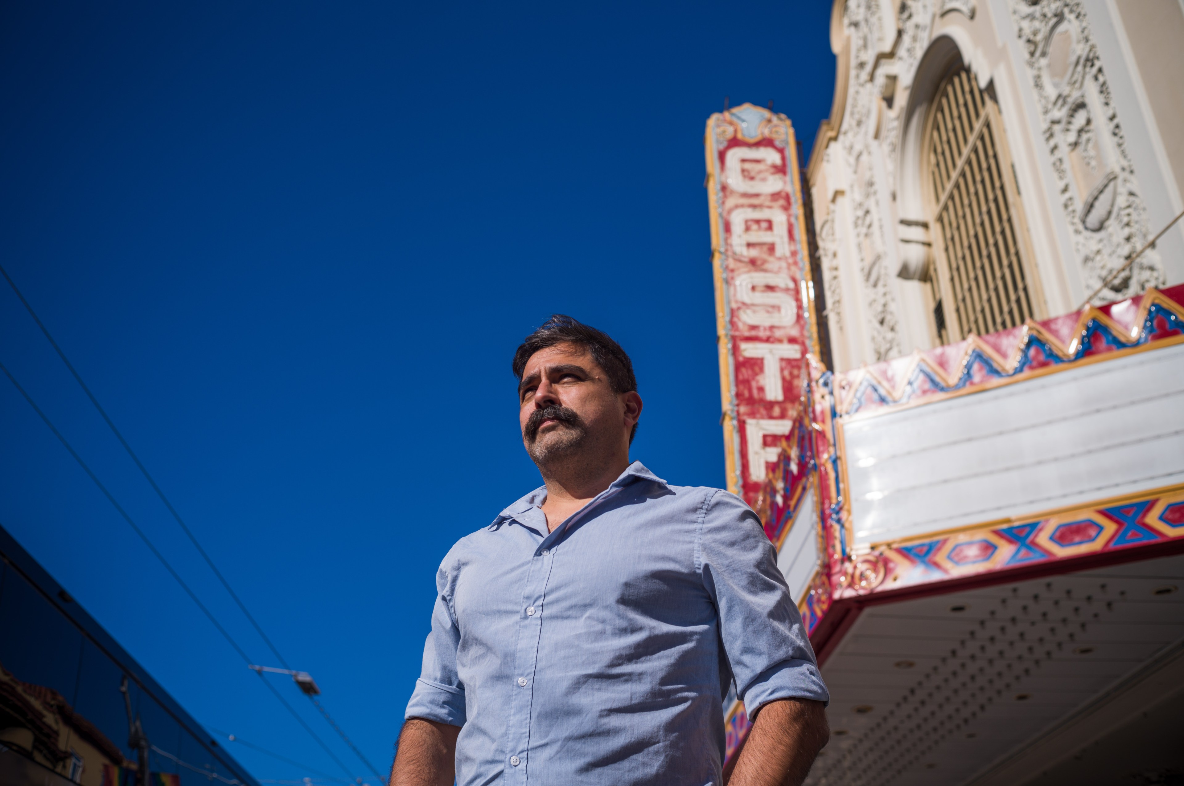 A man stands in front of an ornate theater marquee under a clear blue sky.