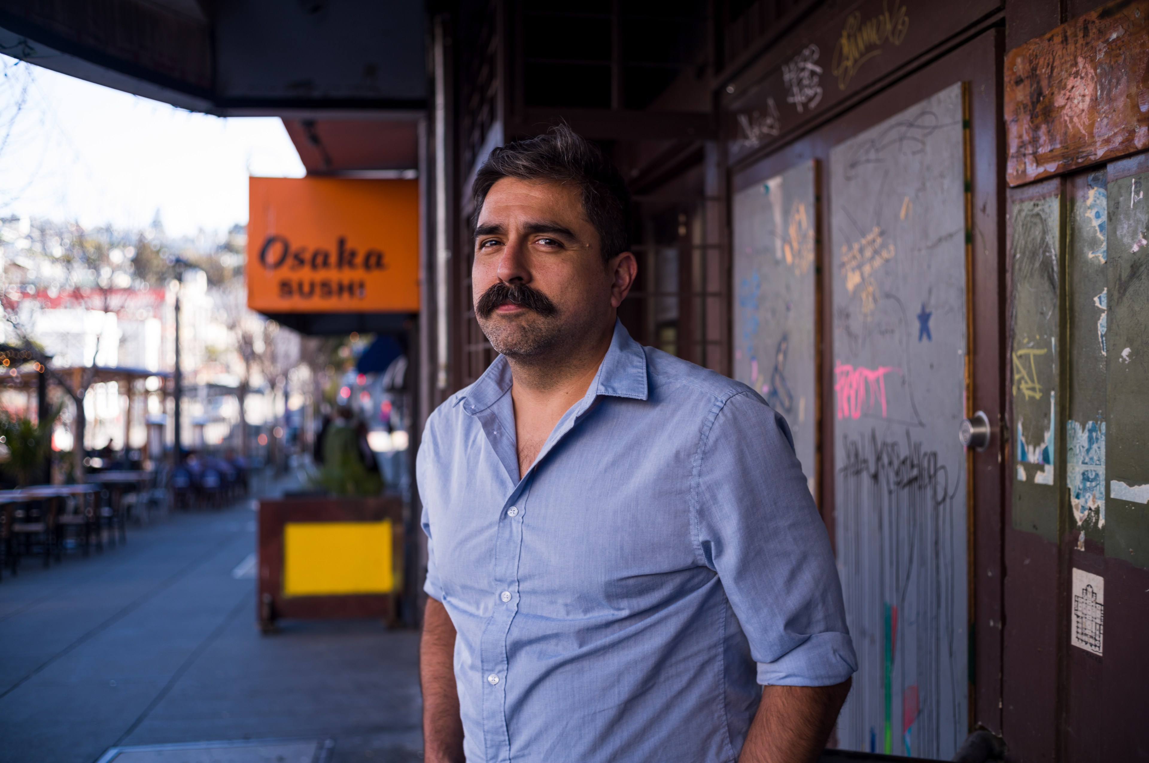 A man with a mustache stands by a graffiti-covered wall near &quot;Osaka Sushi&quot; sign on a sunny street.