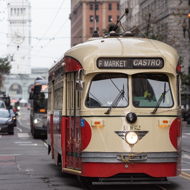 A vintage streetcar in cream and red colors marked "F Market Castro" travels on urban tracks, with a blurred city backdrop.