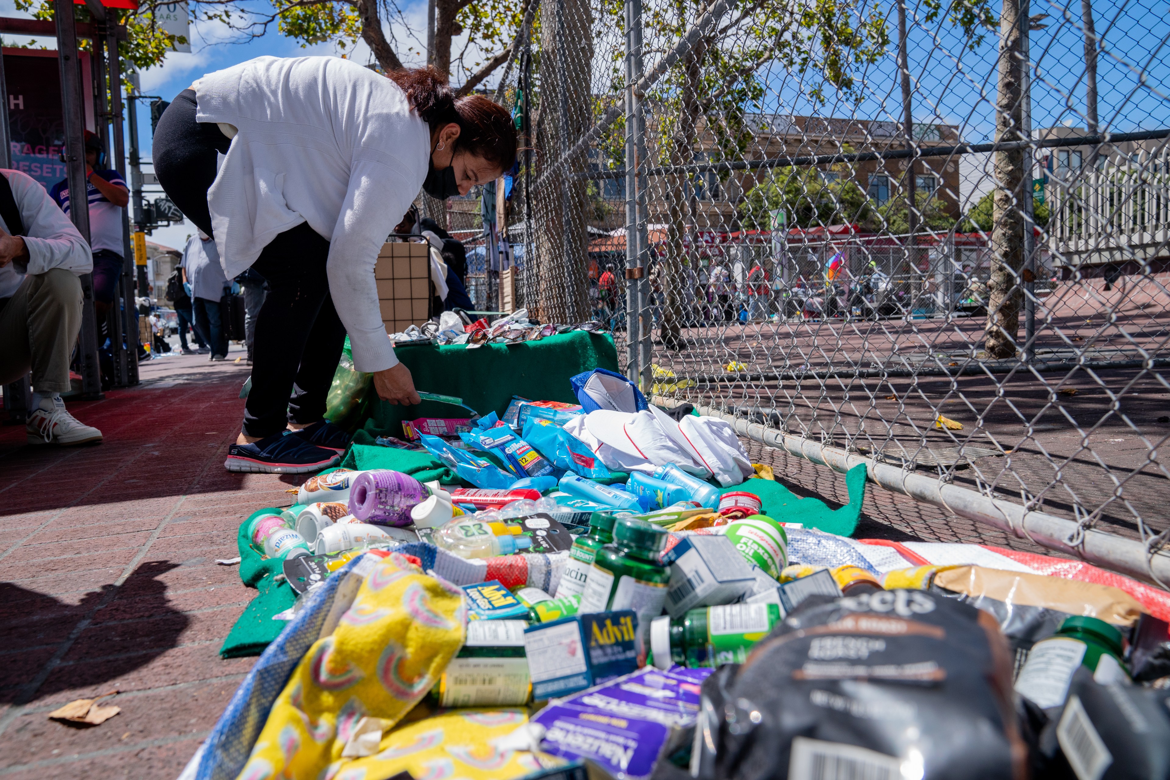 A person is sifting through various items including Advil, coffee, wipes, and laundry detergent along Mission Street in San Francisco, with people walking by in the background.