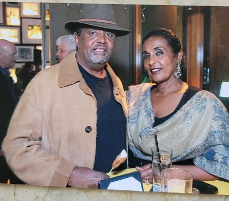A man in a tan jacket and brown fedora stands beside a woman with a scarf draped over her shoulders.