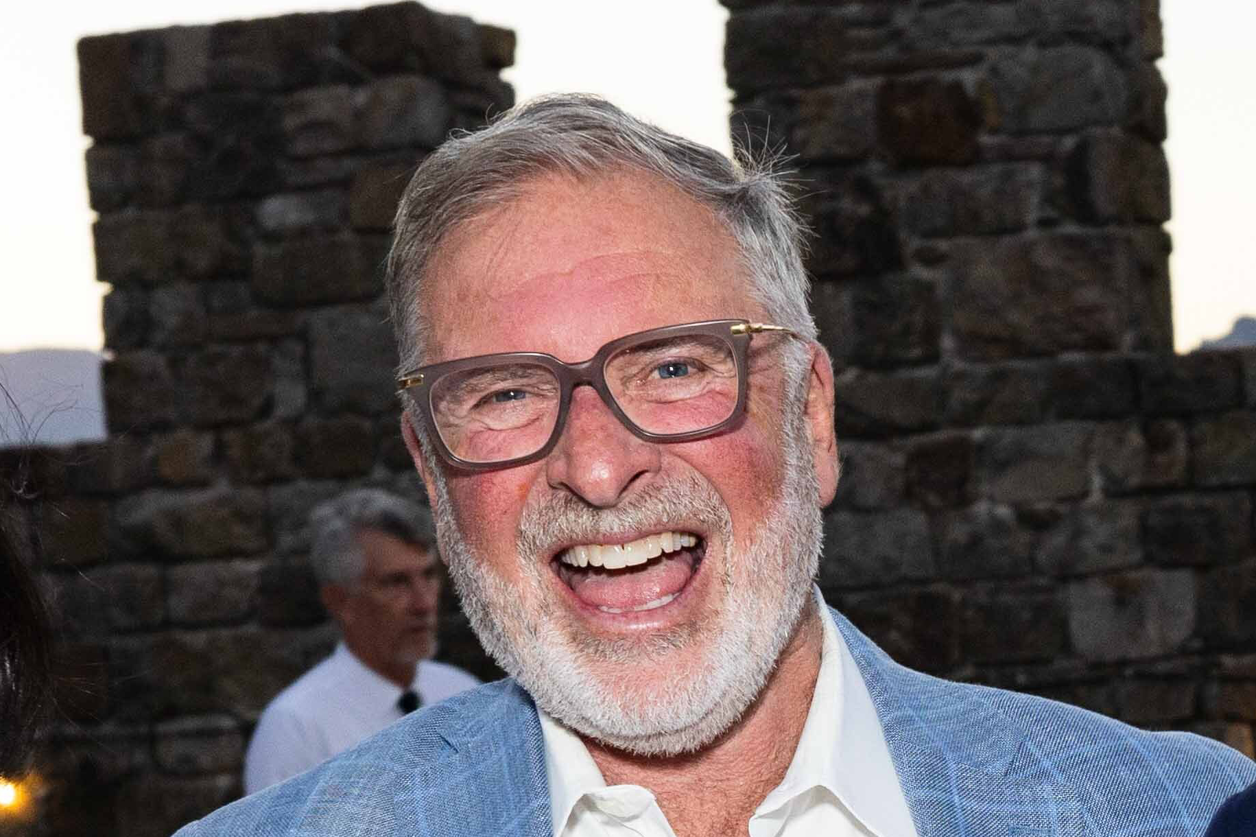 A man with a thin beard, while wearing a blue sport coat laughs out loud with brown stone in the background.