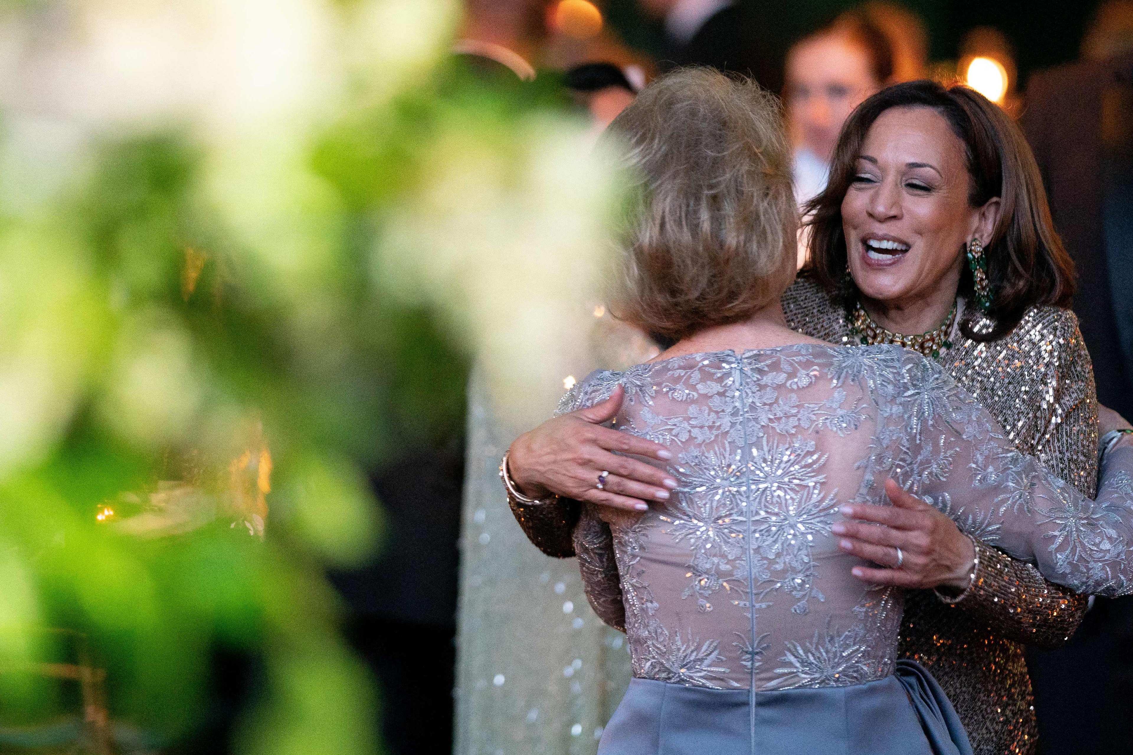 US Vice President Kamala Harris wearing formal wear smiles while she hugs an unidentifiable woman, also wearing formal wear, in a formal dining room setting with an out of focus flower arrangment in the foreground.