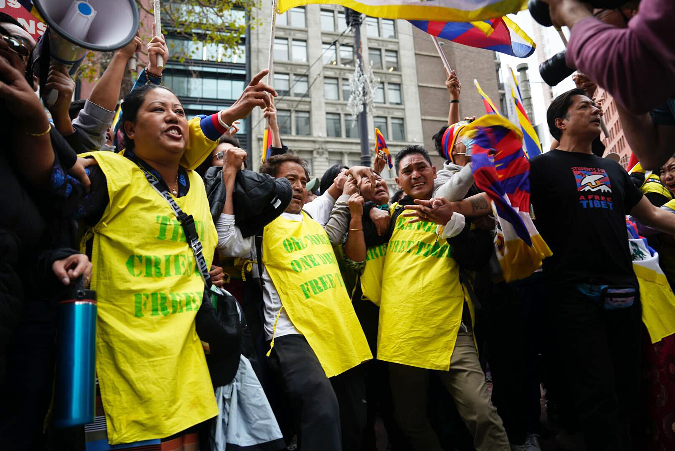A group of people link arms during a protest with Tibetan flags in the background.