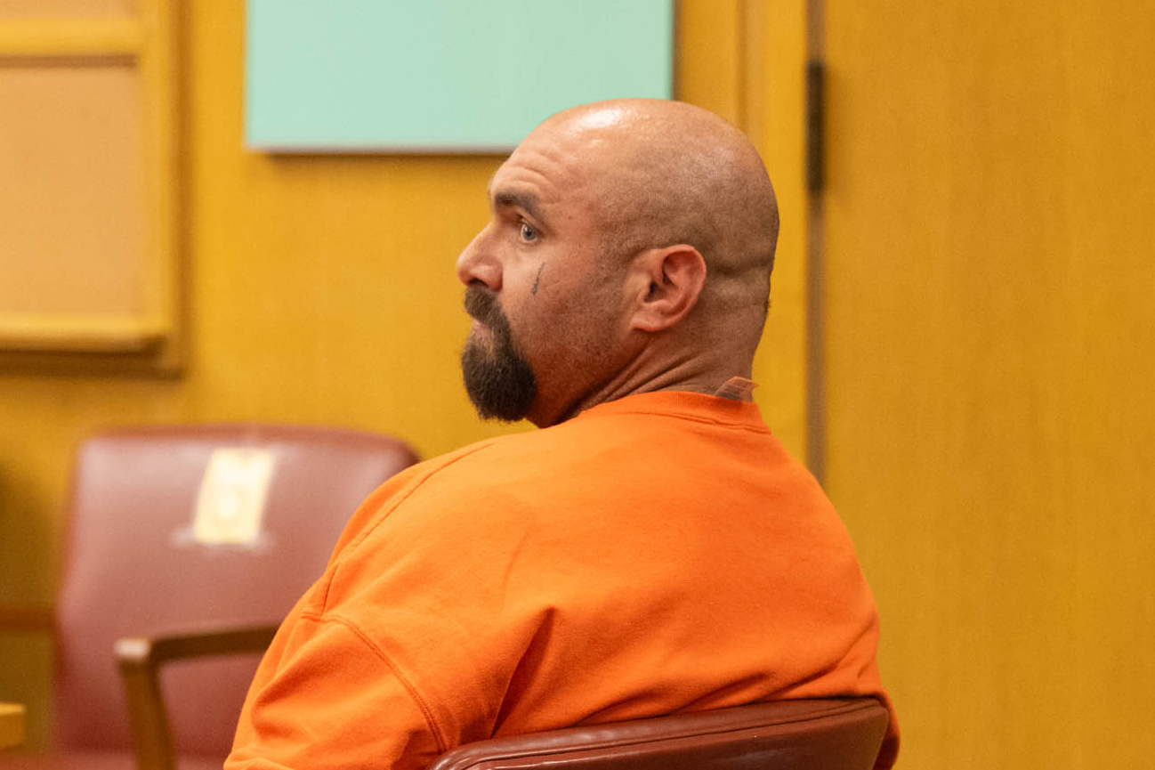A shaven-headed goateed man in an orange shirt is seen from his left side as he sits in a courtroom.