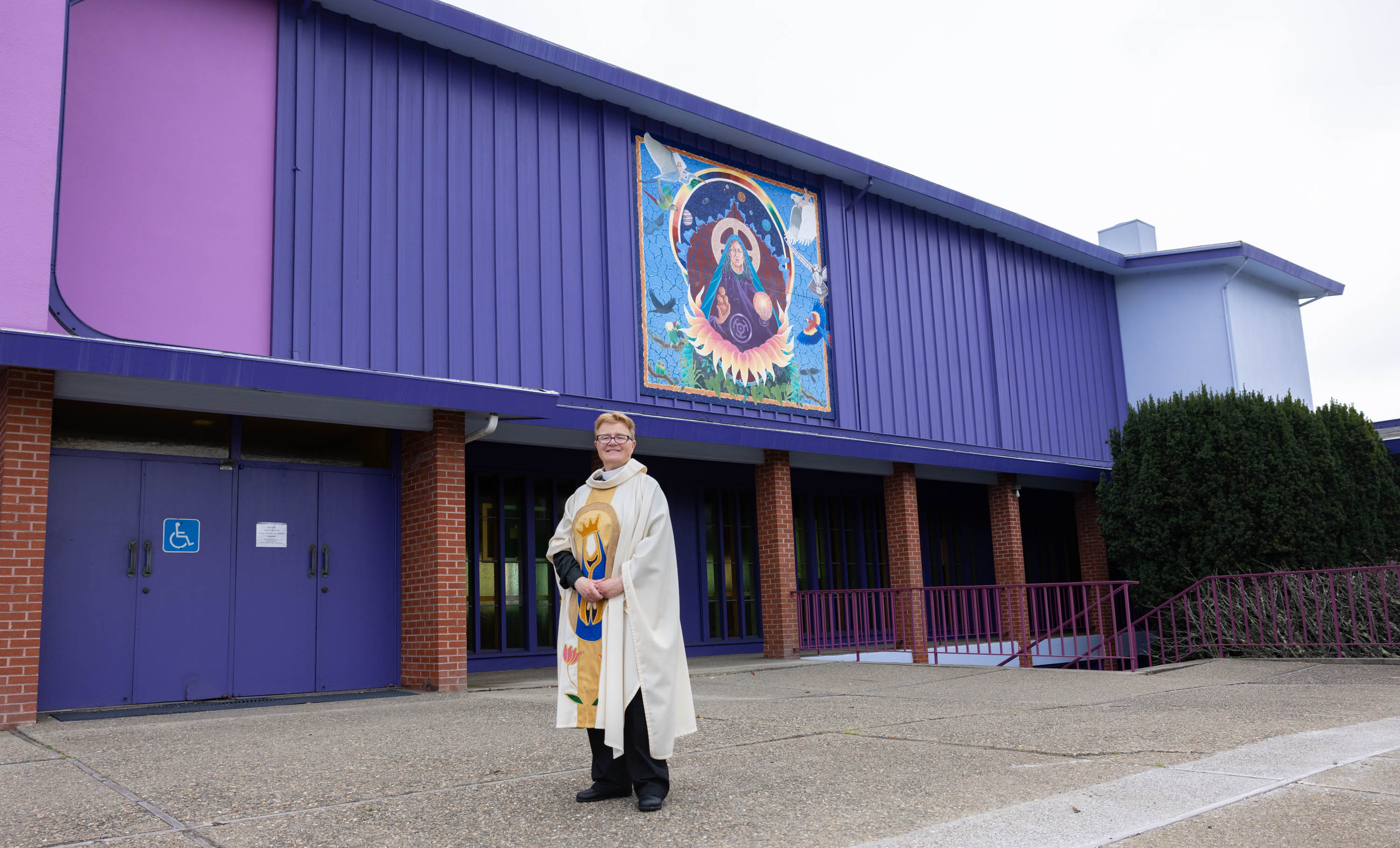 A pastors stands in front of the exteririor of a large purple church