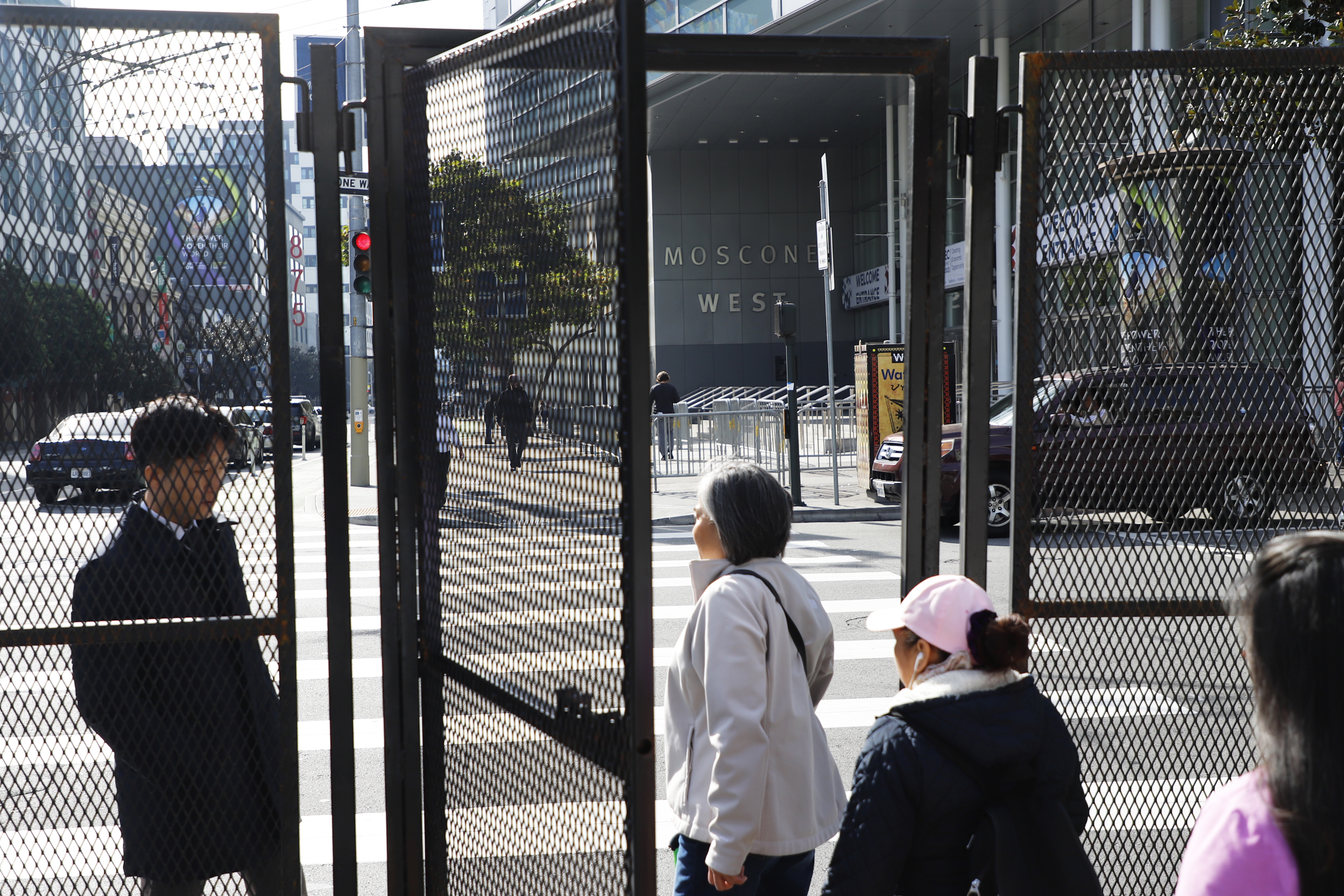 A person walks through a tall black metal fence with an open gate are on a city street corner with a building that reads "Moscone West" in the background.
