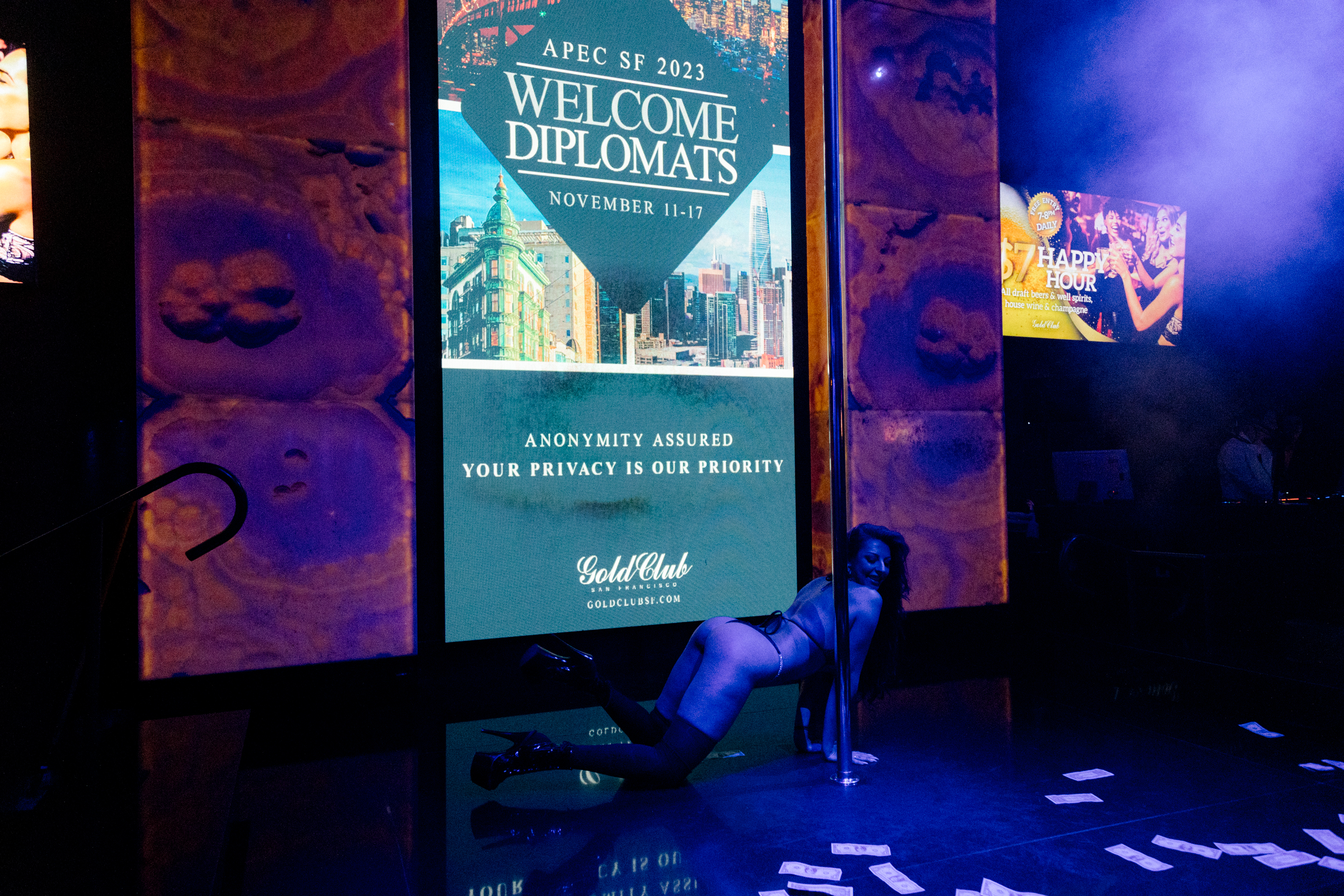 An exotic dancers is on her hands and knees in the middle of the dance floor with dollar bills on the ground and a LCD screen that reads &quot;Welcome Diplomats - APEC SF 2023 - November 11-17&quot;