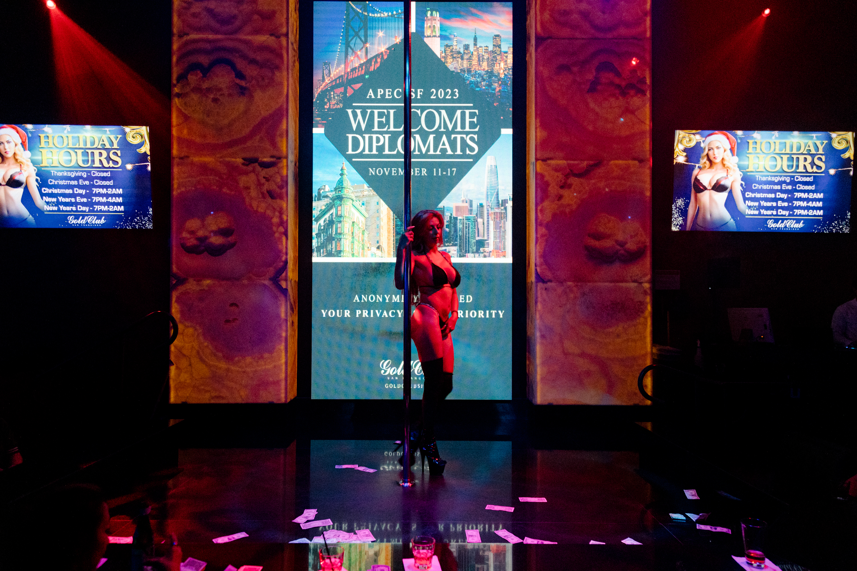 An exotic dancers stands in the middle of the dance floor with dollar bills on the ground and a LCD screen that reads "Welcome Diplomats - APEC SF 2023 - November 11-17"