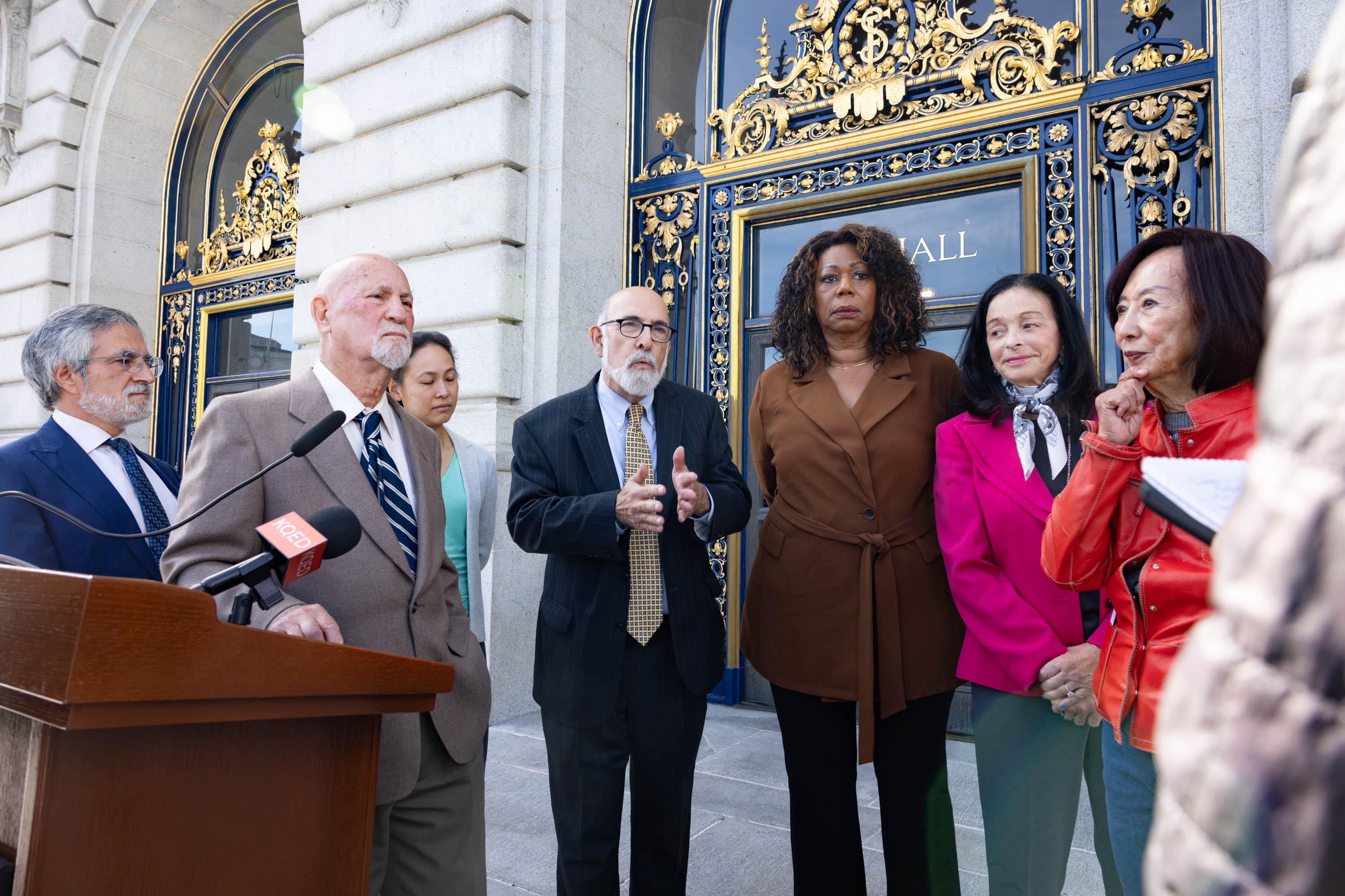 A man in a blue suit gesticulates while standing next to group of people stand im front of a gilded facade of San Francisco's City Hall.