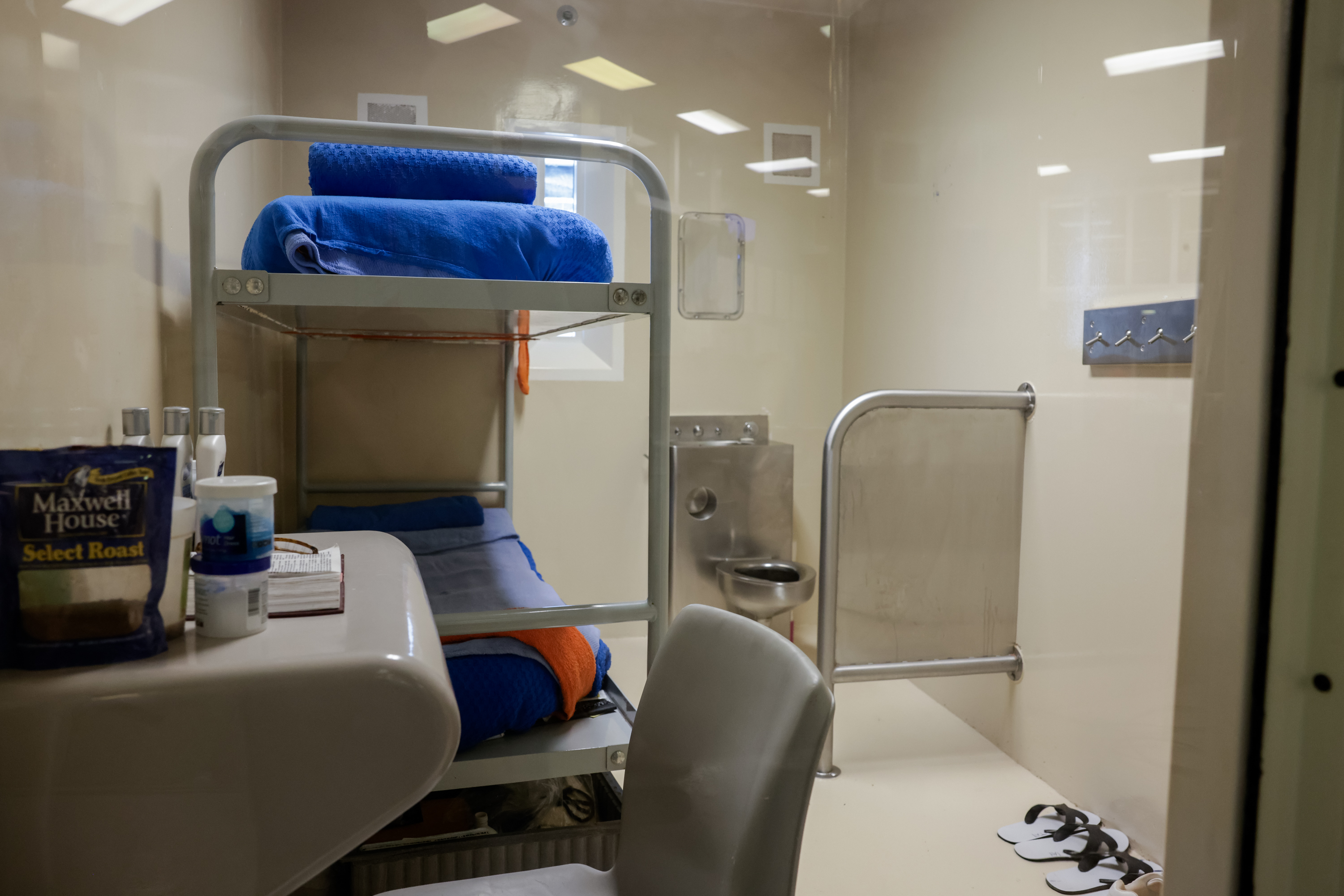 The setting is inside a jail cell which contains two empty beds, a desk, and a toilet.