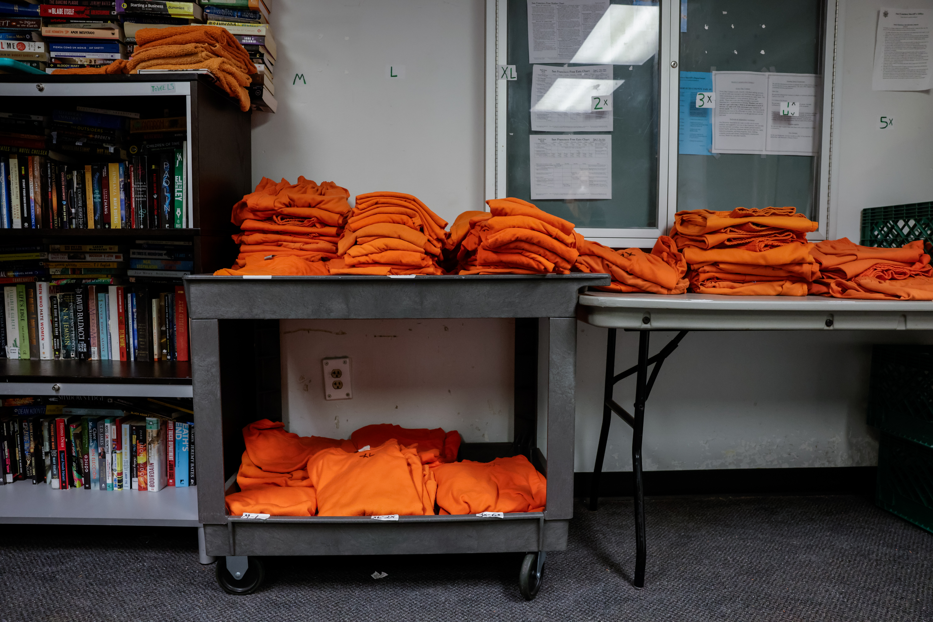 A stack of orange prison clothes sit atop a cart and folding table next to a bookshelf inside the jail.