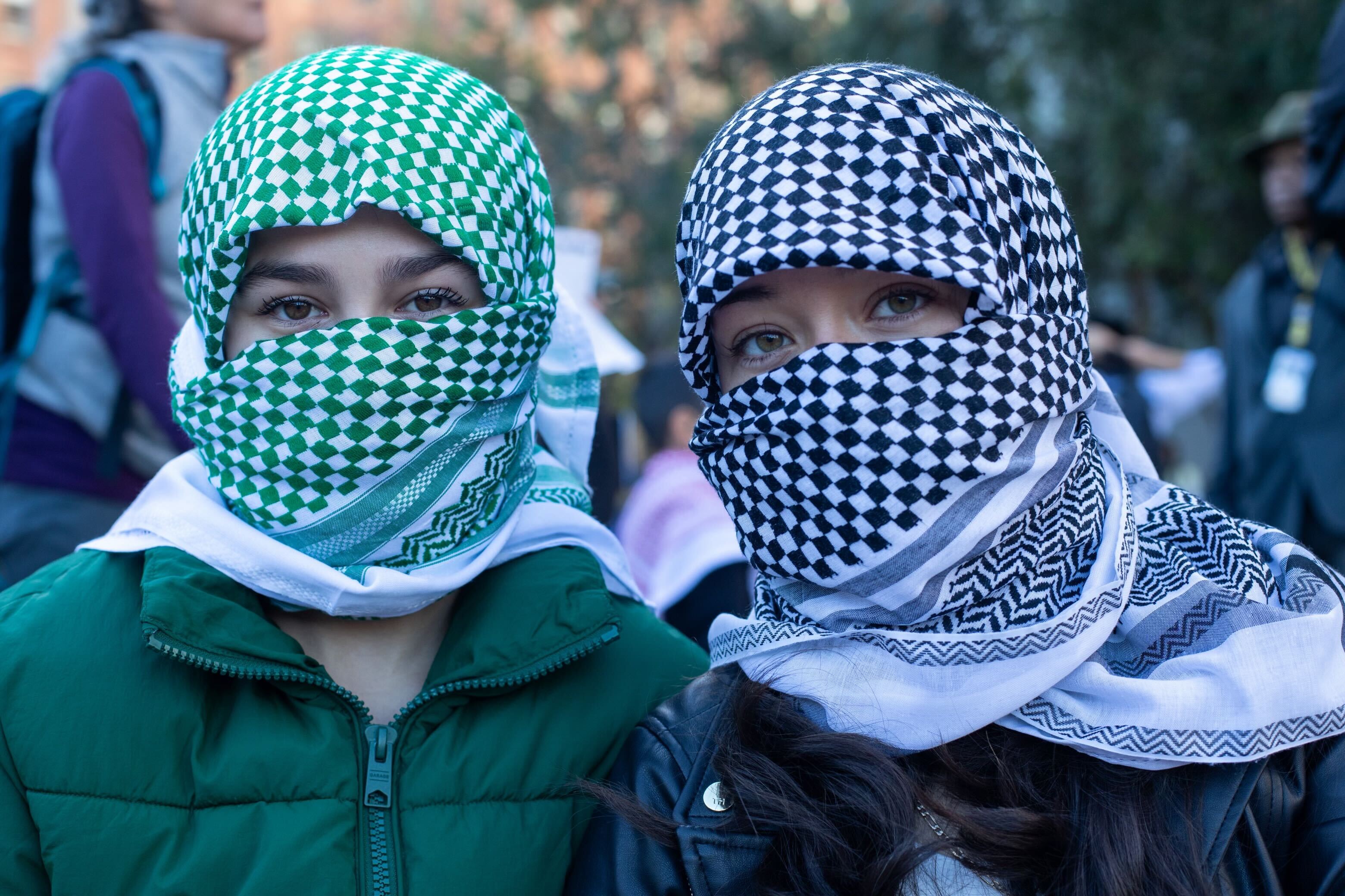 Two women wearing keffiyeh scarves look at a photographer taking a photo of them.